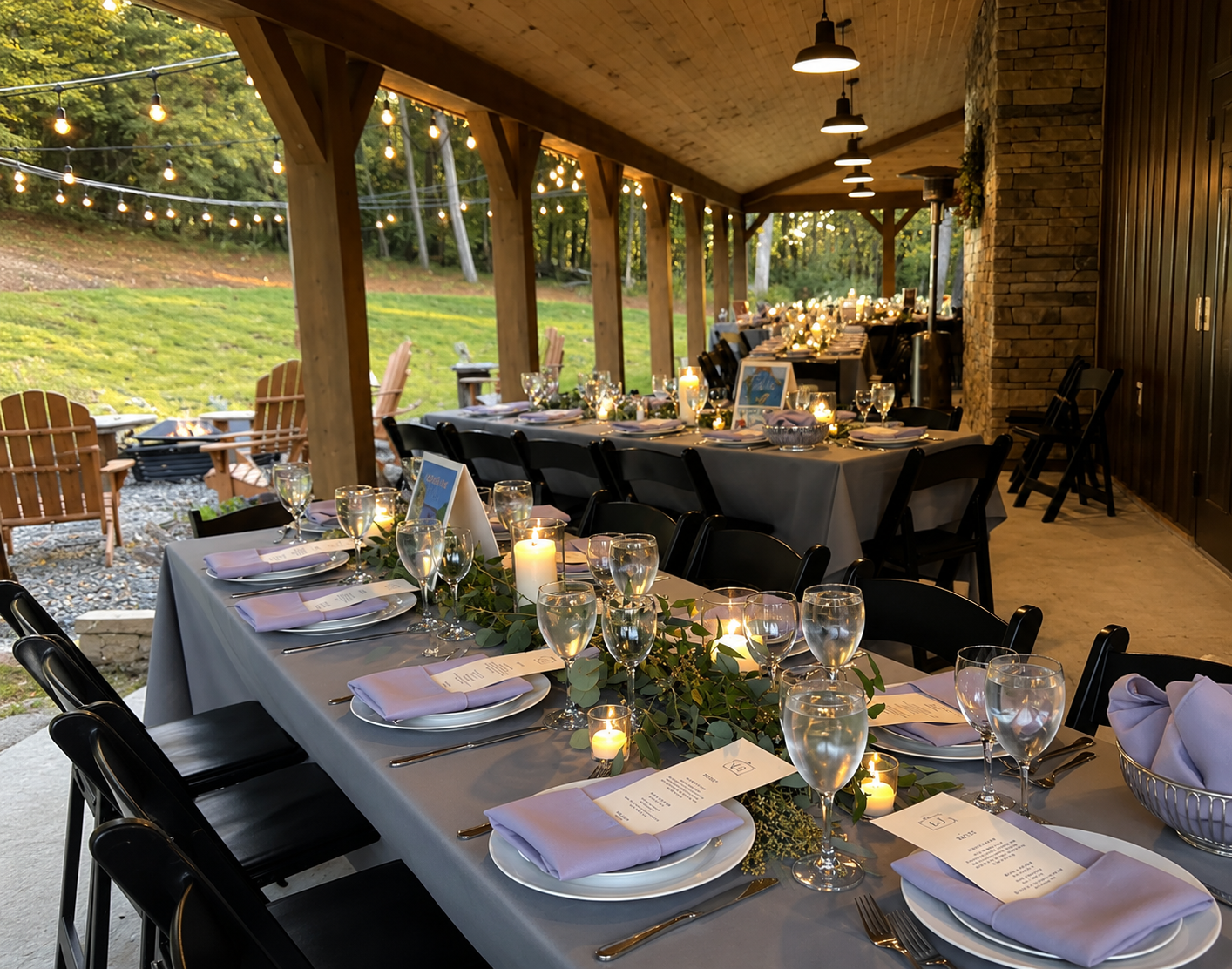 A decorated outdoor dining area with tables set for an event, featuring string lights, candles, wine glasses, and place settings, with a grassy yard and outdoor chairs in the background.