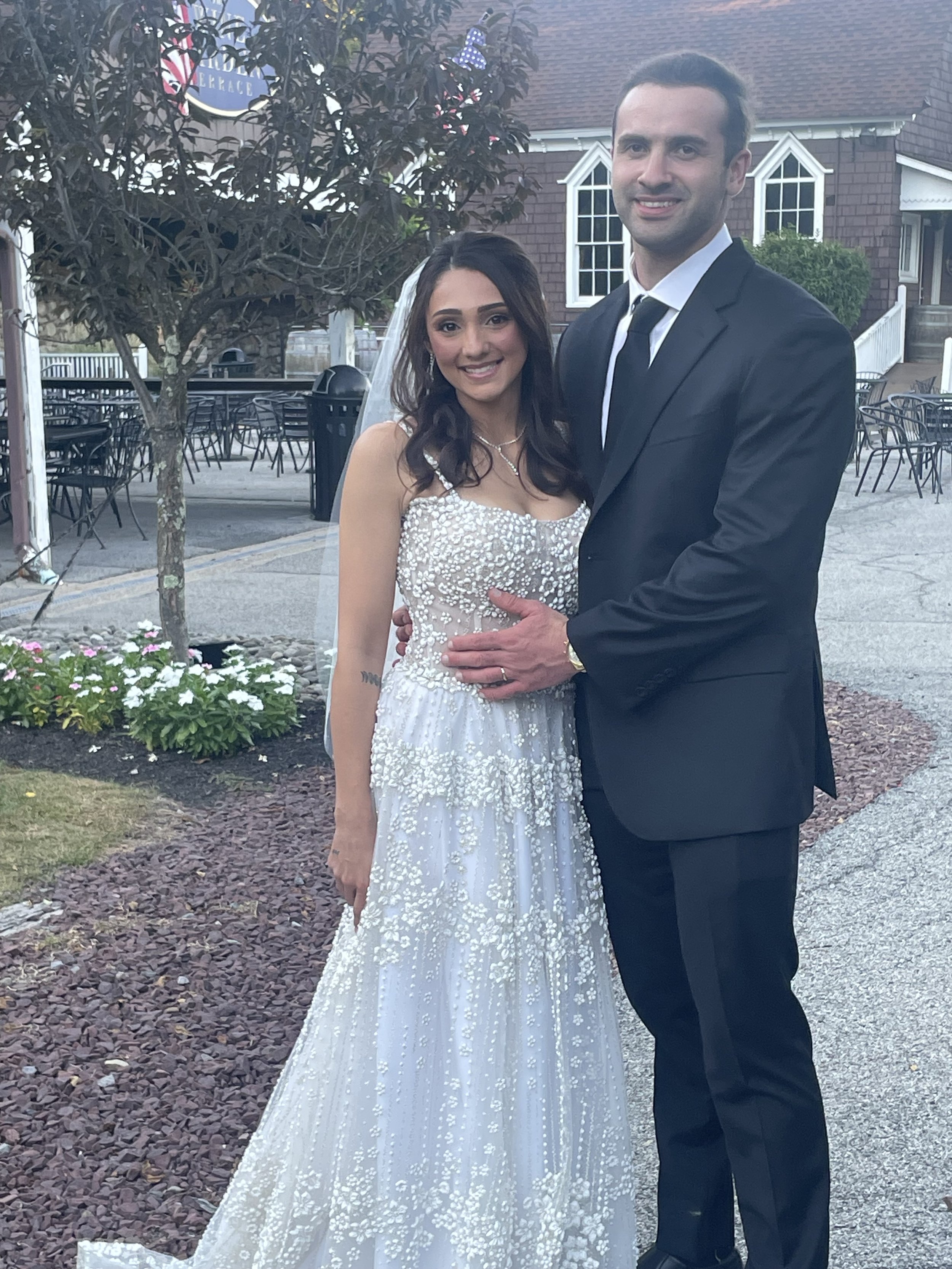 A couple wearing wedding attire stands outdoors in front of a red brick building, with the bride in a white dress and veil and the groom in a black tuxedo, smiling and looking at the camera.