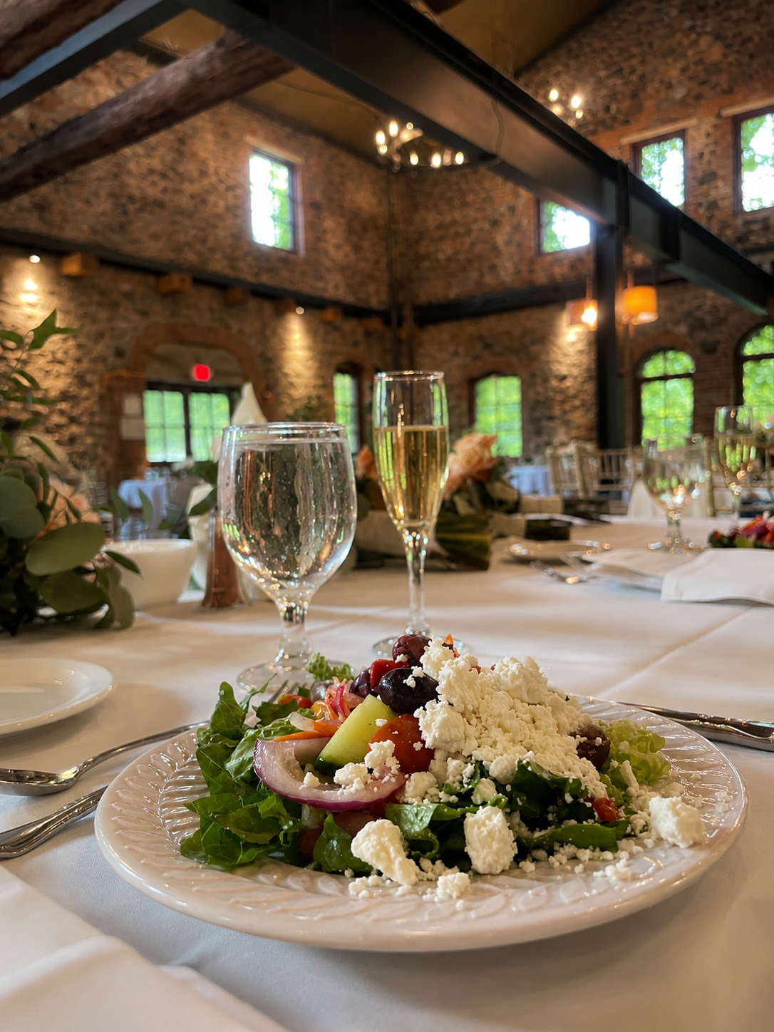 A plate of Greek salad with feta cheese, tomatoes, cucumbers, olives, and red onions on a formal dining table in a rustic restaurant with brick walls, large windows, and hanging lights.
