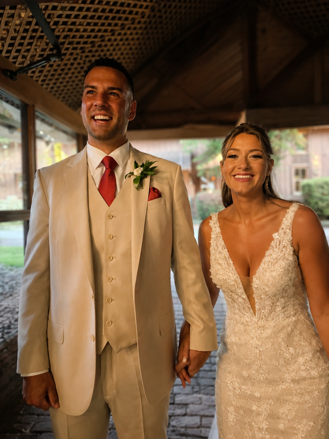 A man and woman dressed in wedding attire, holding hands and smiling indoors with wooden beams and windows in the background.