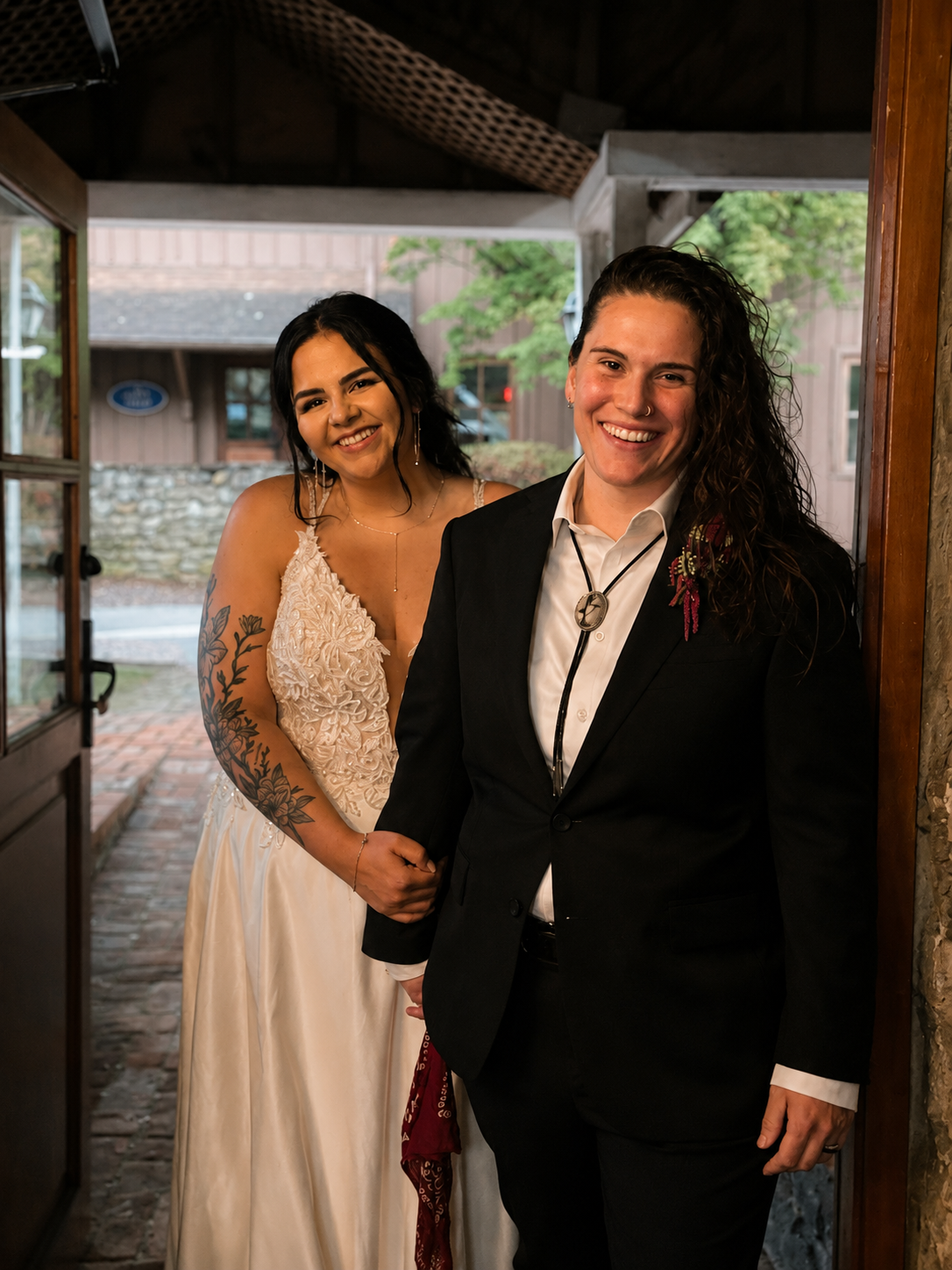 A happy couple at their wedding, standing indoors near an open door, with the woman in a cream dress with lace details and tattoos on her arm, and the man in a black suit with a white shirt and a bolo tie, smiling at the camera.