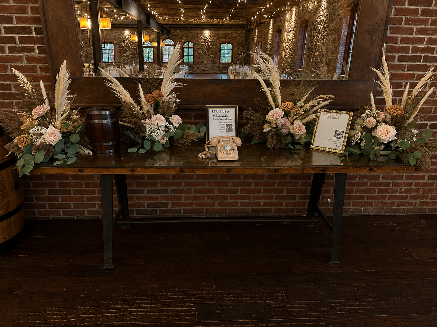 Wedding guest book display with floral arrangements, a vintage telephone, and framed signs inside a rustic venue with brick walls and wooden flooring.