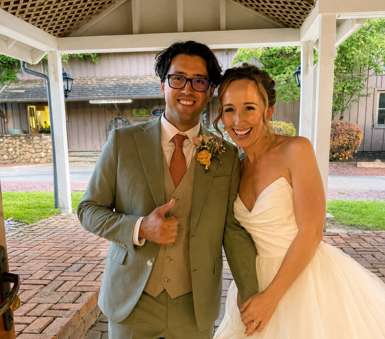 A smiling bride and groom posing together outside under a pavilion, with the groom giving a thumbs up. The groom is wearing a light gray suit and glasses, with a boutonniere on his lapel. The bride is in a strapless white wedding dress, with her hair