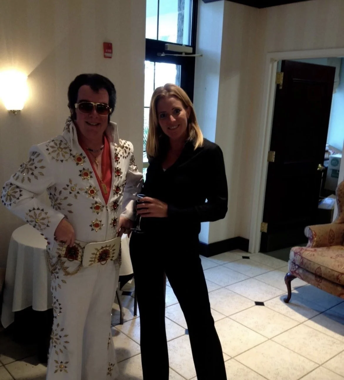 Two women standing together indoors, one dressed as Elvis Presley in a white jumpsuit with gold and red embellishments, and the other in a black outfit holding a glass of wine, smiling at the camera.
