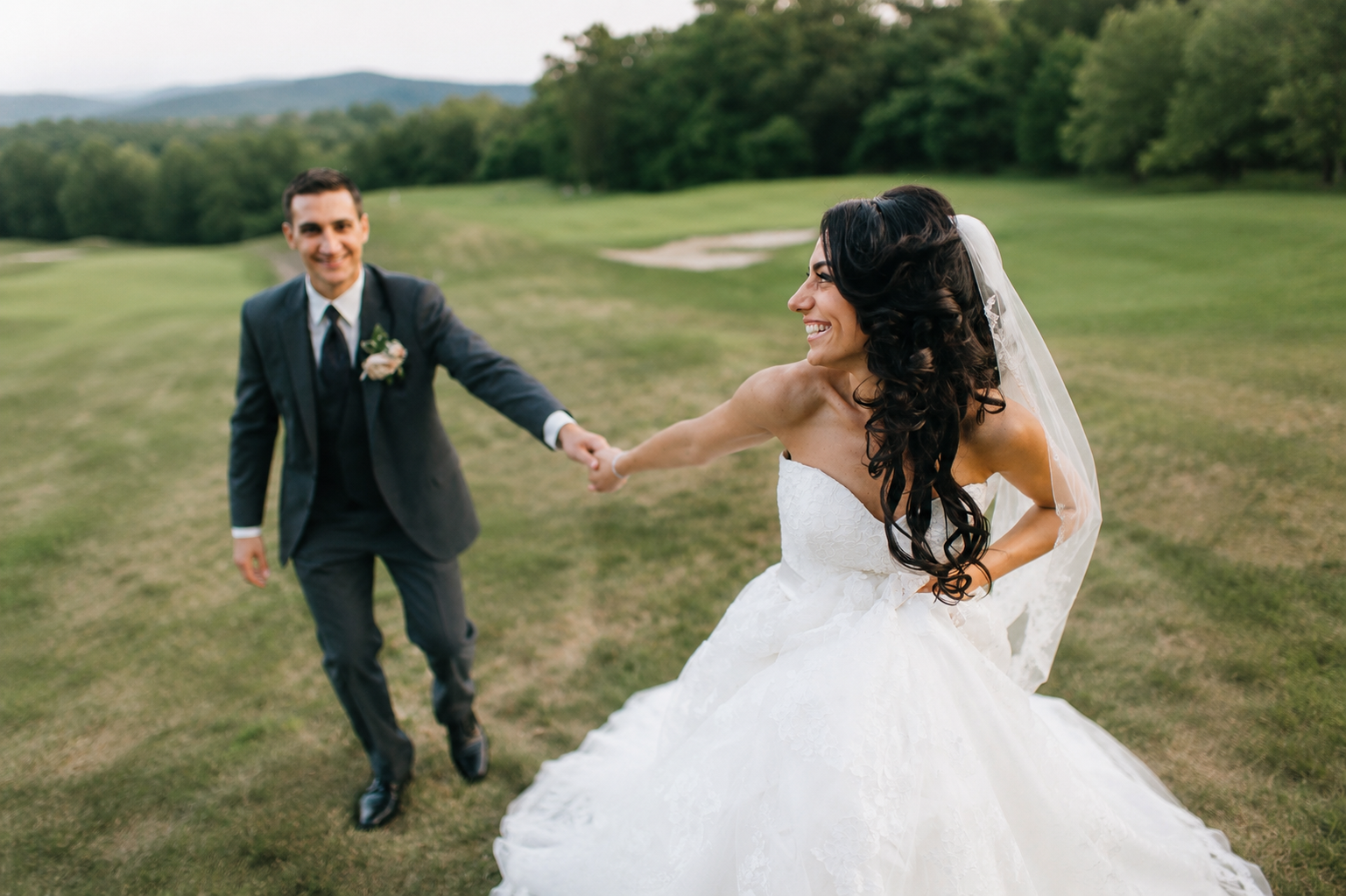 A smiling bride with dark, curly hair in a white wedding gown and veil holding hands with a groomsman in a black suit and tie on a grassy field with trees and hills in the background.