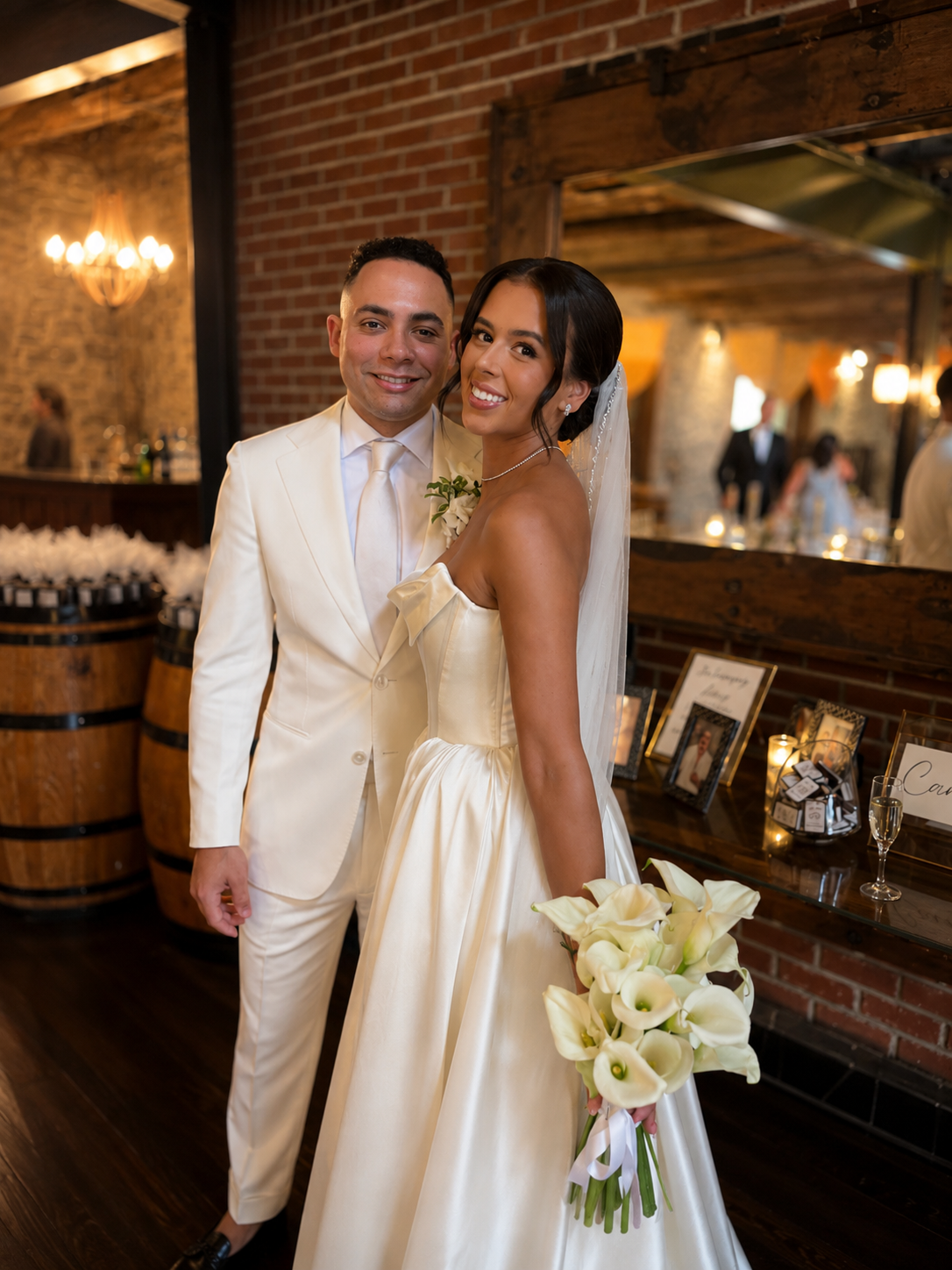 A newlywed couple standing close together inside a rustic wedding venue, smiling at the camera. The groom is wearing a white tuxedo, and the bride is wearing a strapless white wedding gown holding a bouquet of calla lilies. The background features br