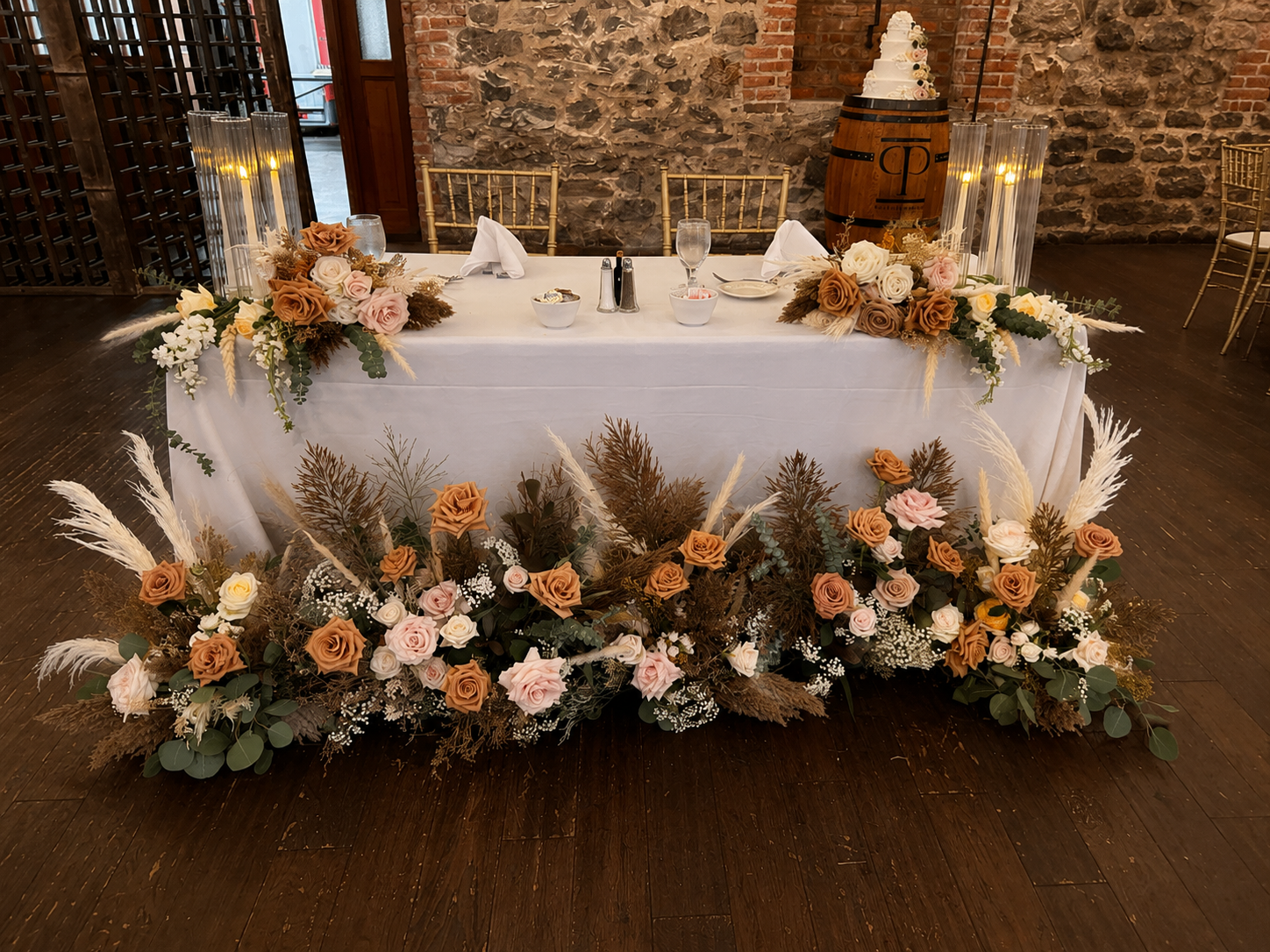 A wedding reception table decorated with flowers and surrounded by wooden chairs. A decorative arrangement of flowers and foliage extends along the front of the table, with a large barrel and wedding cake in the background.