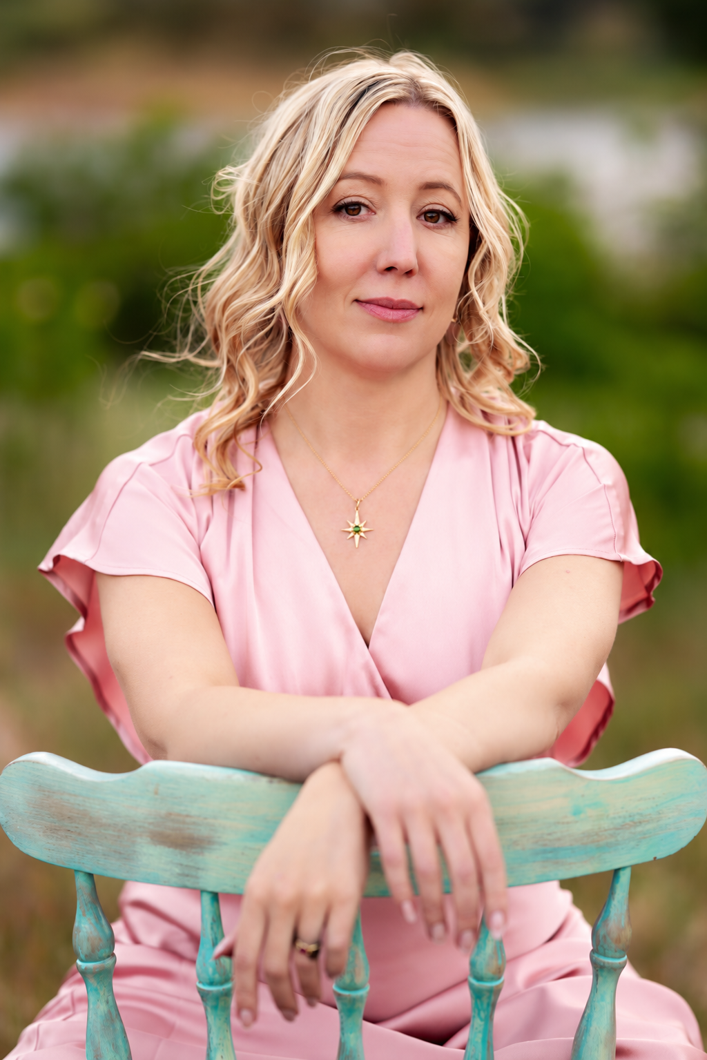 A woman with blonde curly hair wearing a pink dress and a star-shaped necklace, sitting outdoors on a light blue wooden chair, with a blurred green natural background.