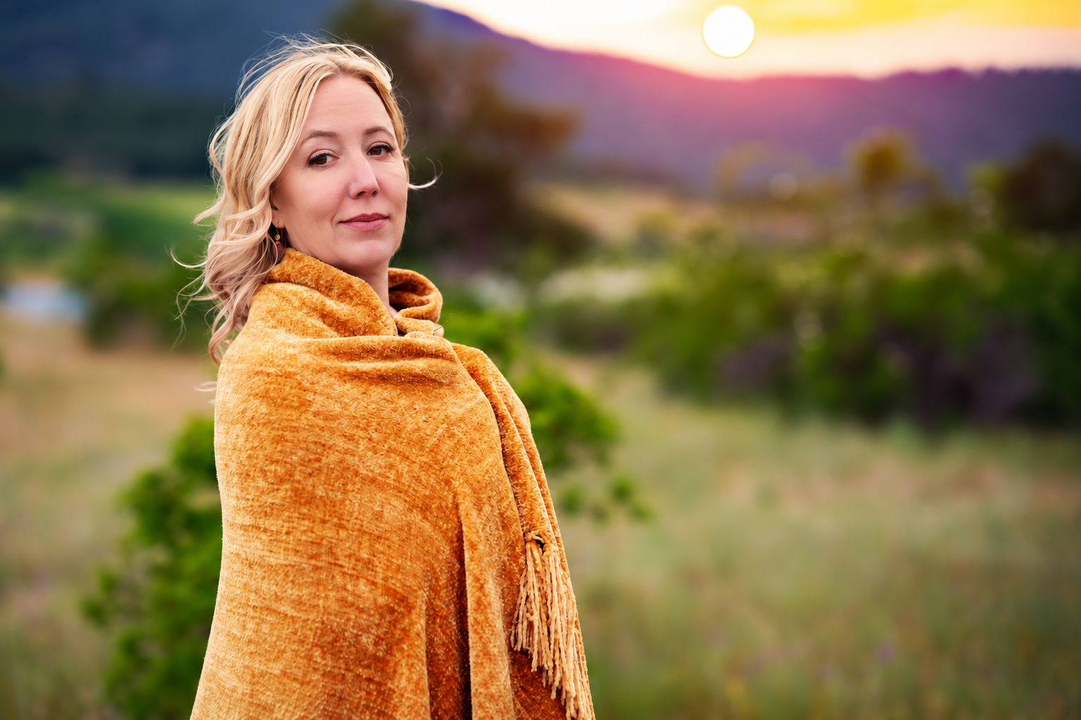 A woman with blonde hair in a loose wave, wearing a cozy orange-brown shawl, outdoors during sunset, with green grass, trees, and a purple mountain in the background.