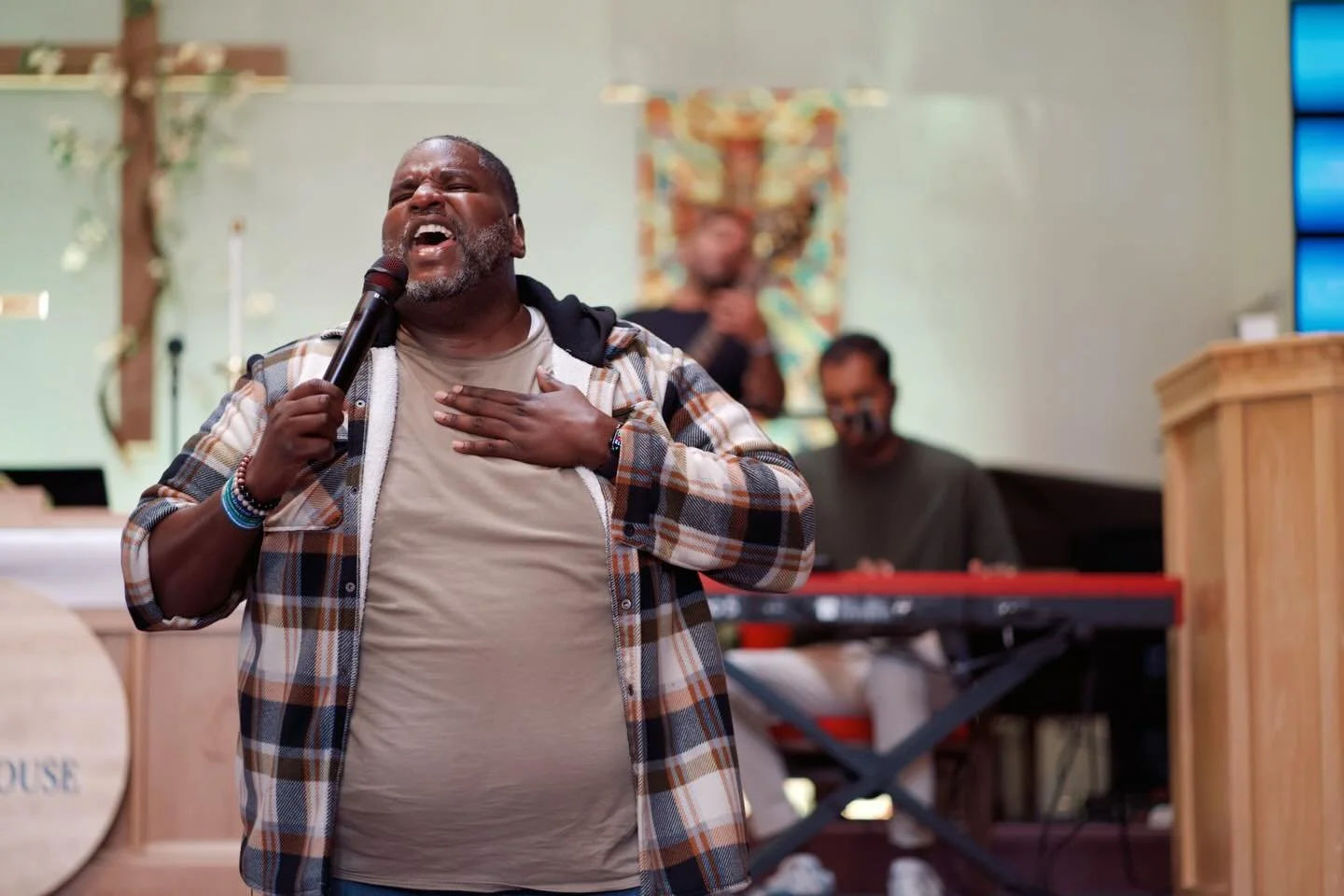 A man singing passionately into a microphone while touching his chest, with two musicians playing instruments in the background.