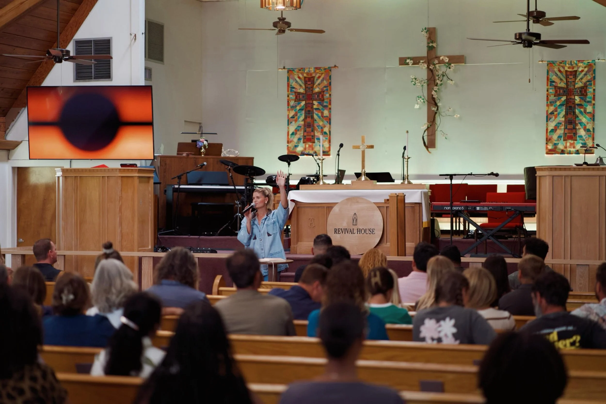 Inside a church with an altar, cross, banners, and musical instruments, a woman is speaking or singing into a microphone in front of an audience.
