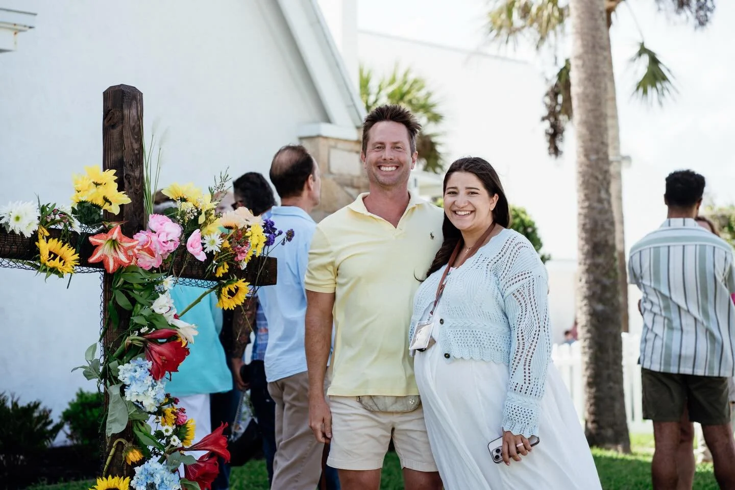 A smiling man and woman standing outdoors, with a floral display in the foreground and palm trees in the background, at a social gathering or outdoor event.