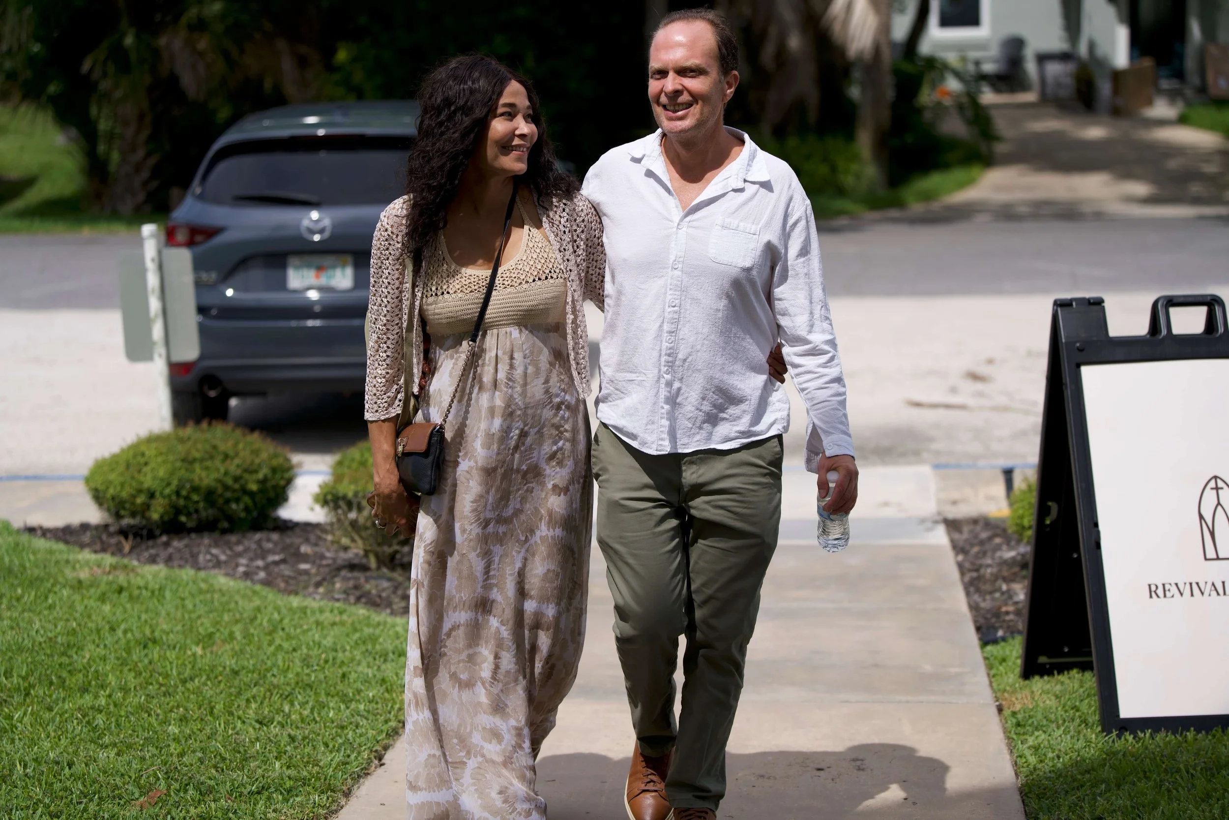 A man and a woman walking outdoors on a sidewalk, smiling and looking at each other. The man is holding a water bottle, and they are near some grass and bushes. There is a black car and a building in the background.