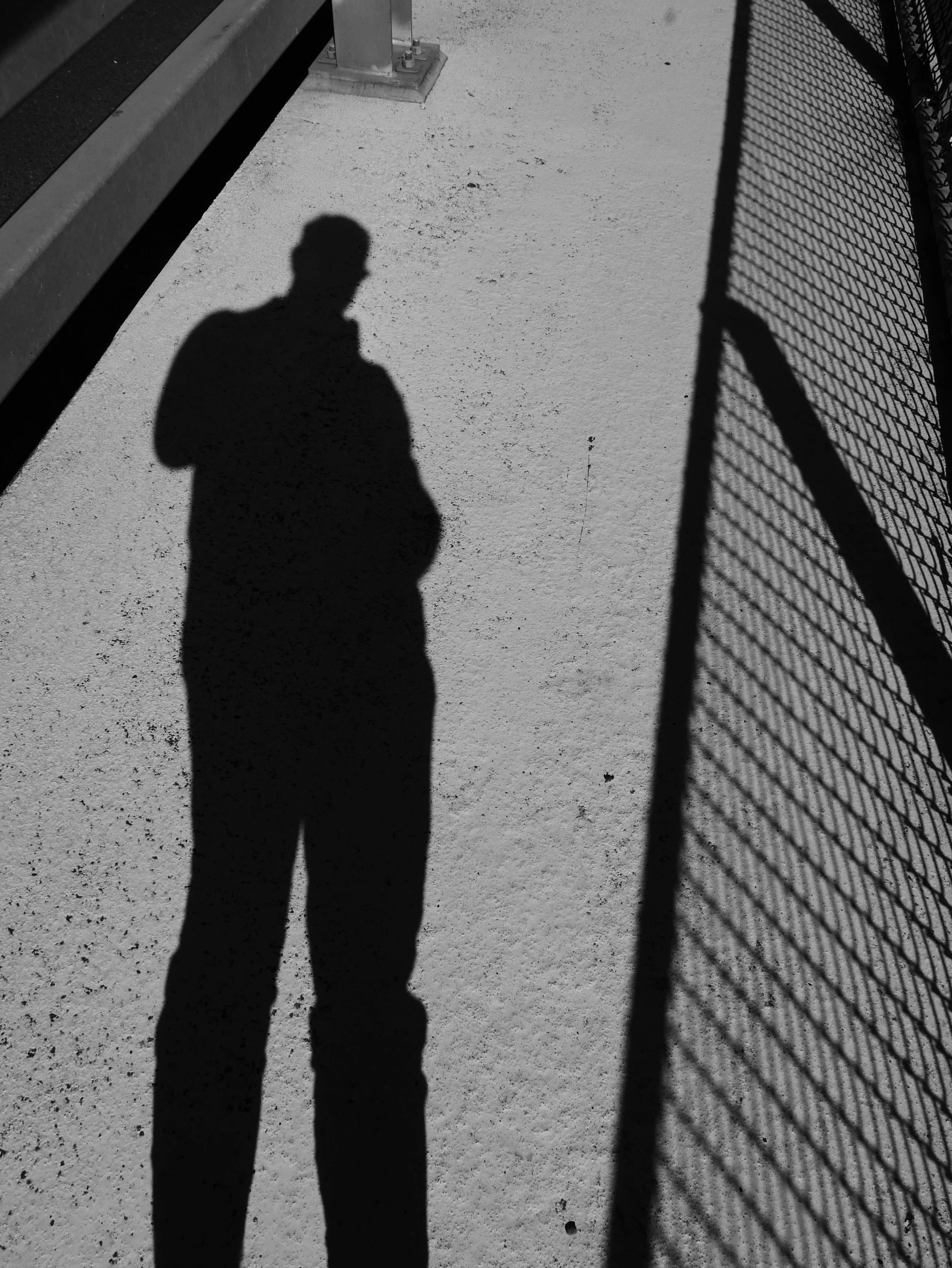 A black and white photo showing the shadow of a person taking a photo, cast on a textured ground. The shadow is next to a chain-link fence, and there is a metal railing and a support structure in the top left corner.