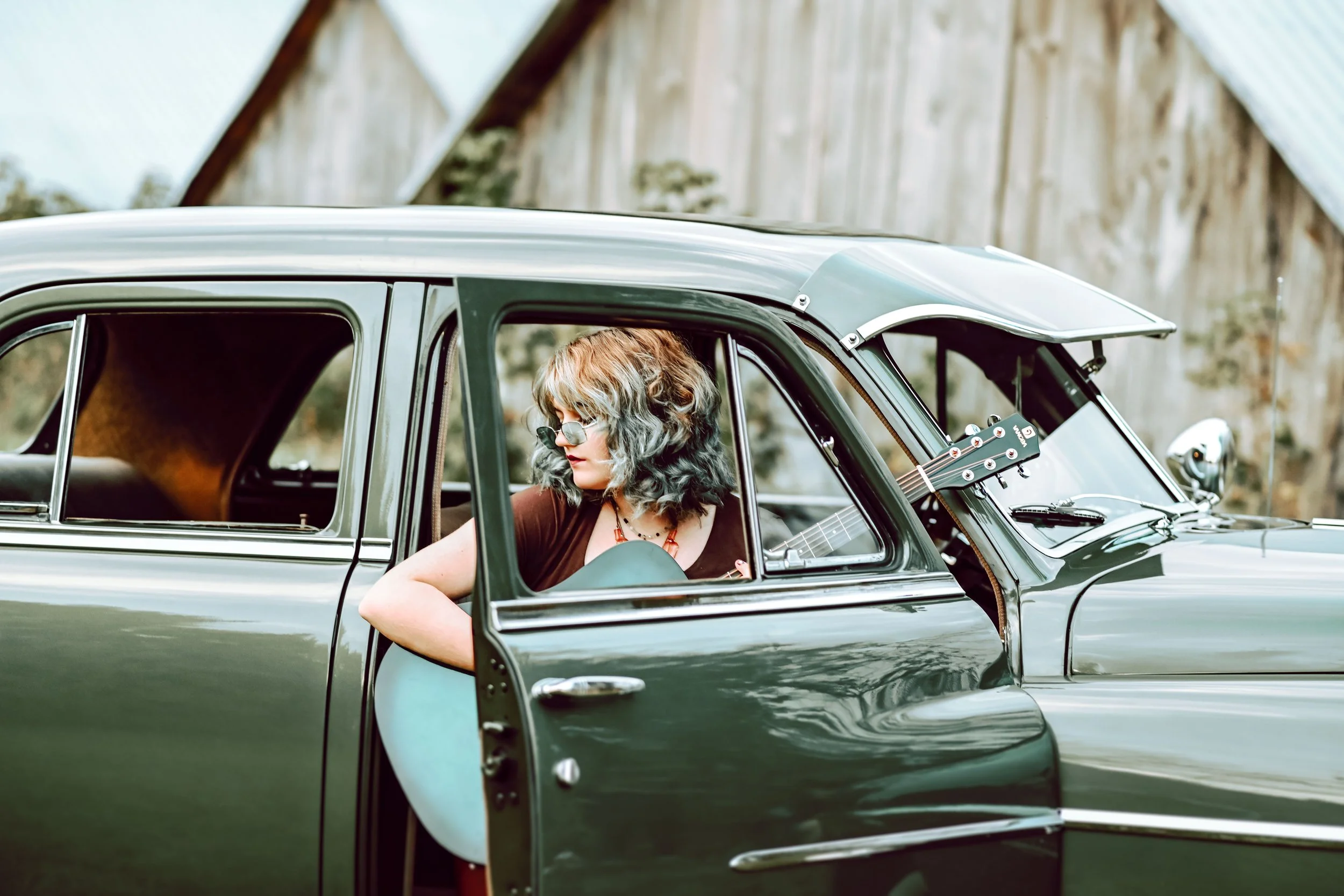 A woman with curly gray hair and glasses sitting in the driver seat of a vintage green car, looking down and playing a guitar, with an outdoor scene and wooden building in the background.