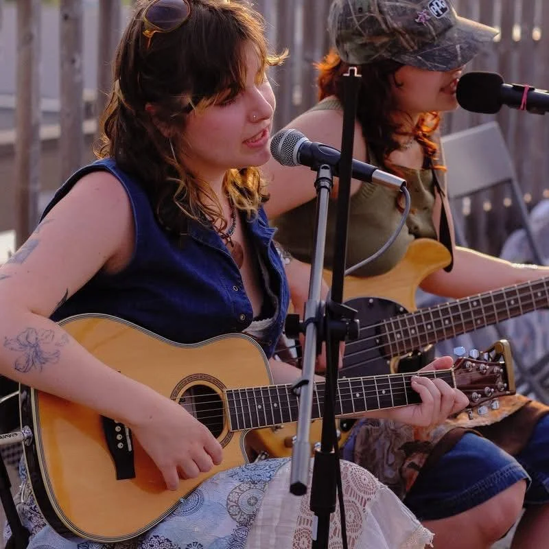 Two women performing music outdoors, one playing an acoustic guitar and singing into a microphone, the other playing an electric bass guitar, both wearing casual clothing.
