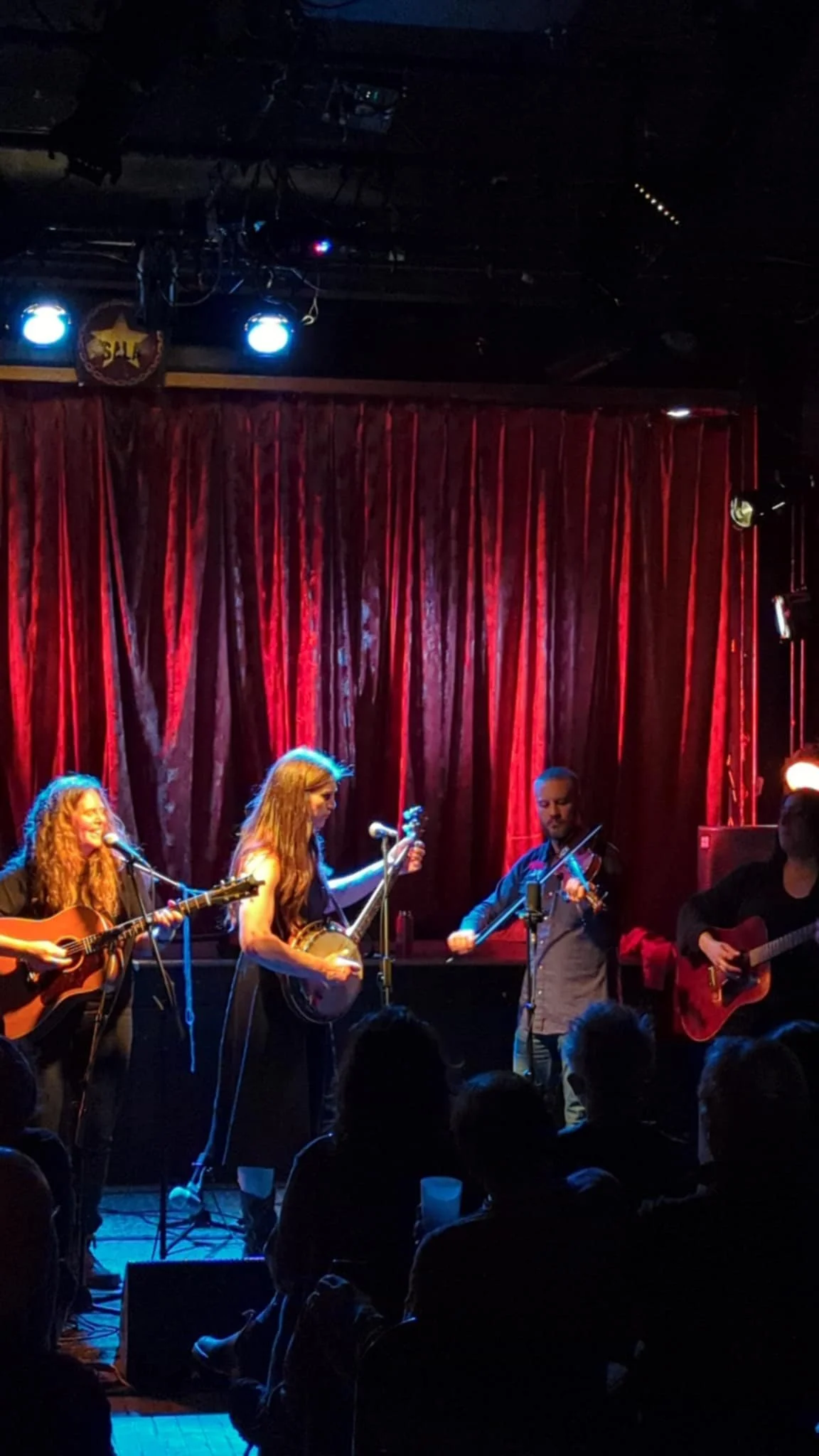 A music performance on stage with four musicians playing instruments, two women and two men, in front of a red curtain, under stage lighting.