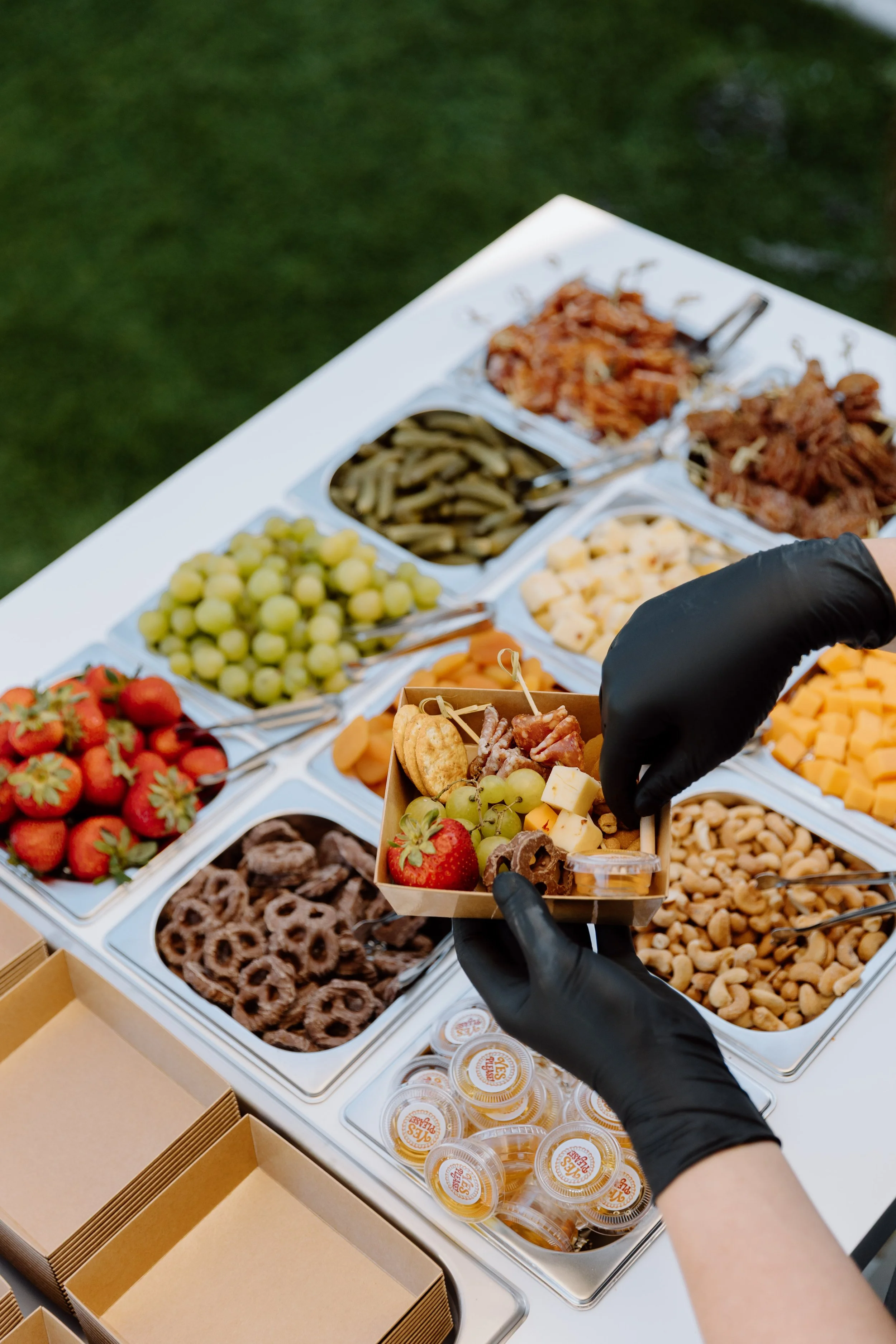 A person wearing black gloves picking cheese and grapes from a buffet table with various snacks and fruits, including strawberries, cookies, and containers of dips, in an outdoor setting.