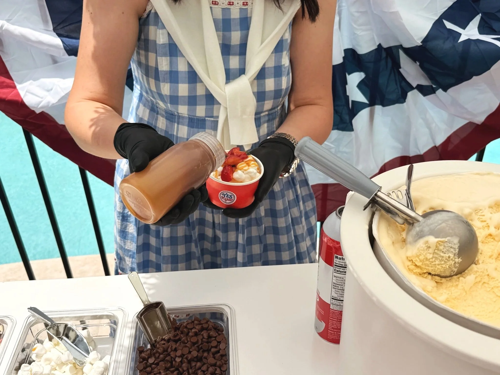 A woman in a blue and white checkered dress is serving ice cream from a large container. She is wearing black gloves and holding a cup with a swirl of ice cream and toppings. There are various toppings and containers of ice cream on a white counter.