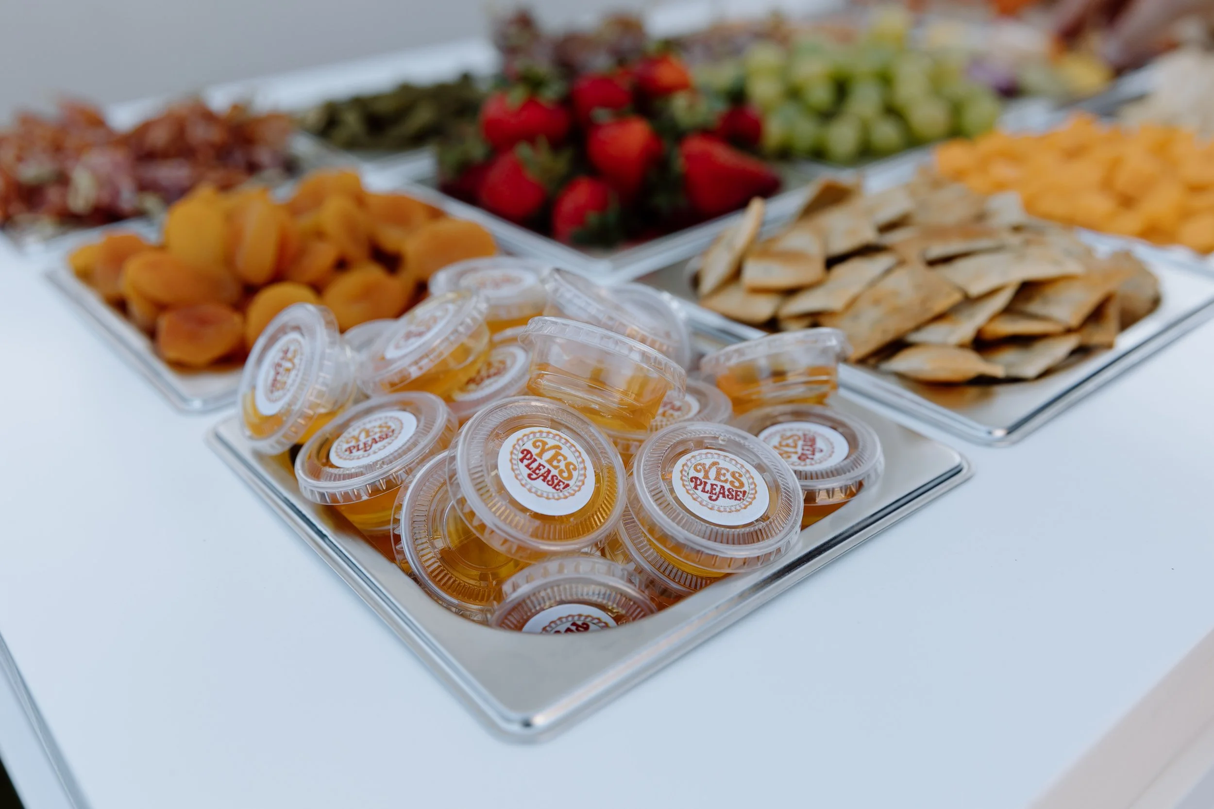 Assorted fruit platter with strawberries, grapes, apricots, and crackers, along with small containers of honey or syrup labeled 'YES PLEASE!'