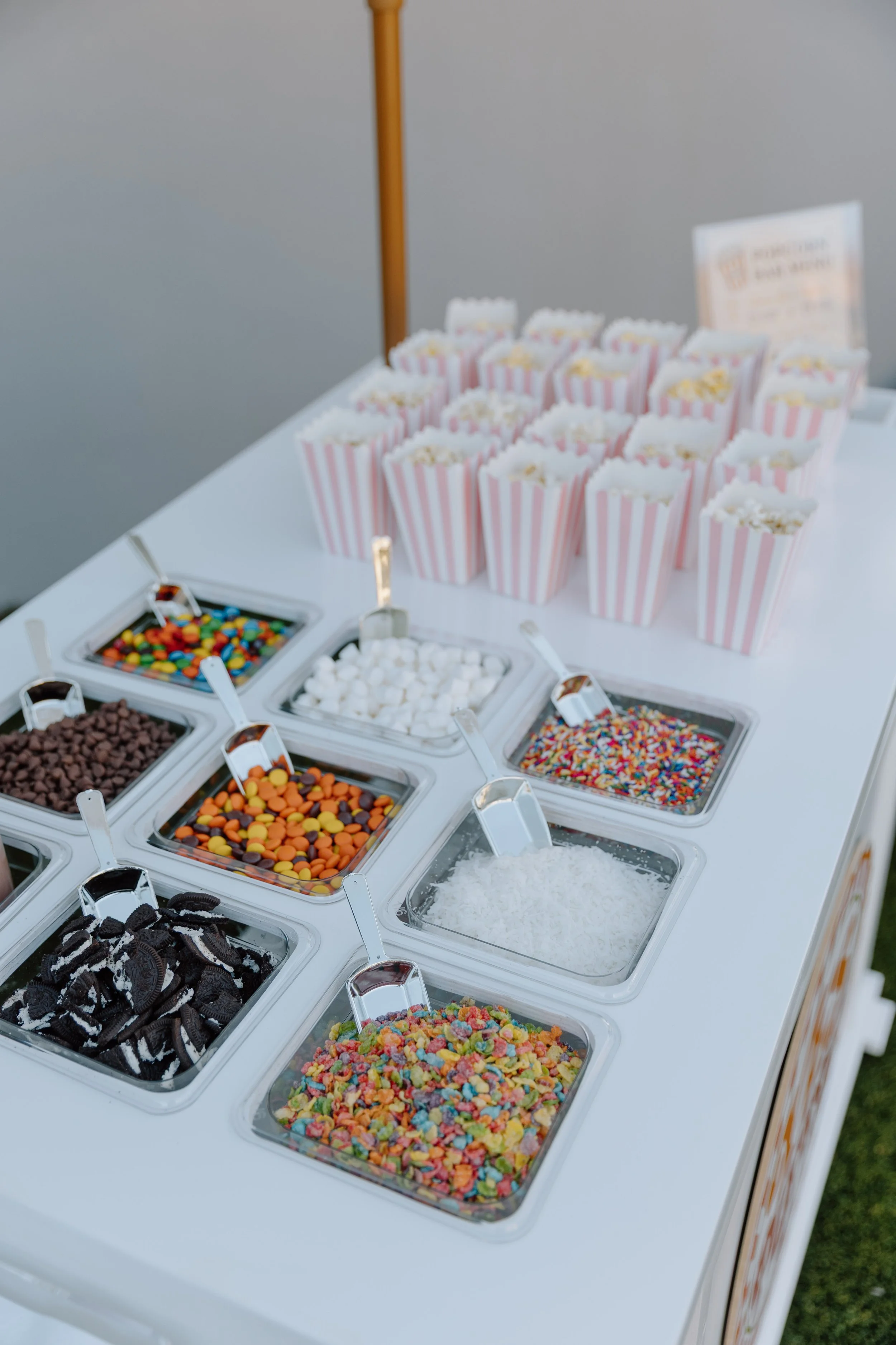 Table with various bowls of candy, including chocolate, gummy bears, sprinkles, and popcorn, with striped popcorn containers in the background.