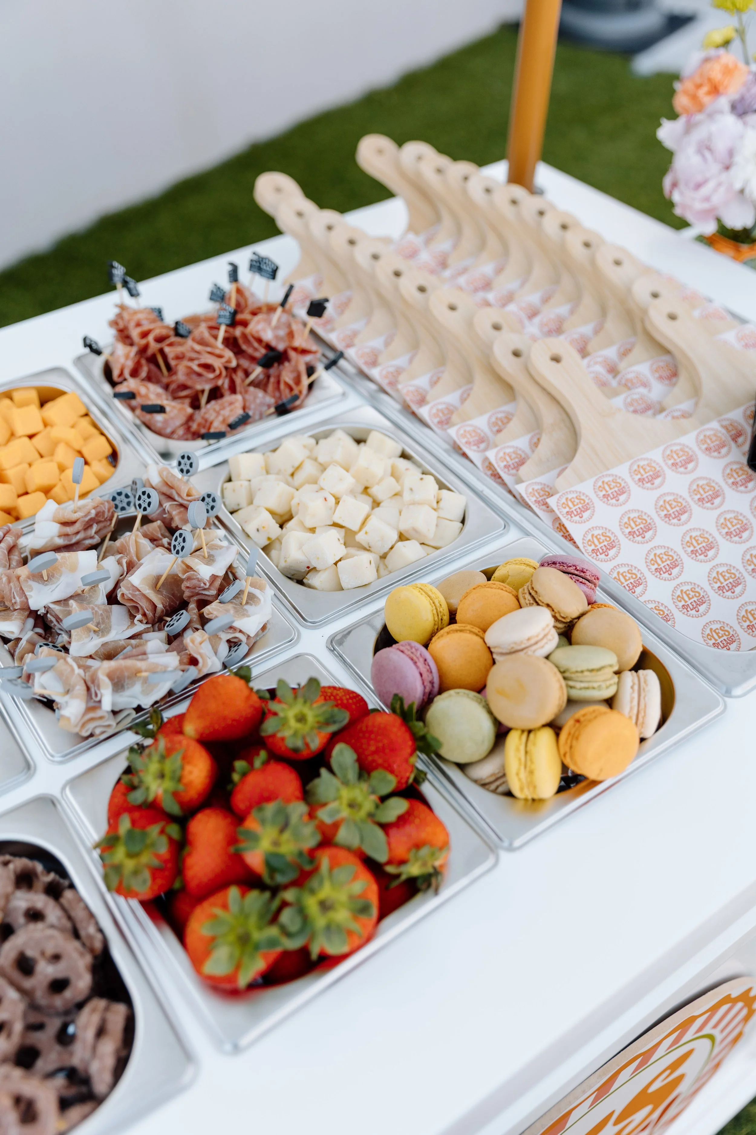 Dessert and snack table with strawberries, macarons, cheese cubes, cured meats, and chocolate cookies at an outdoor event.