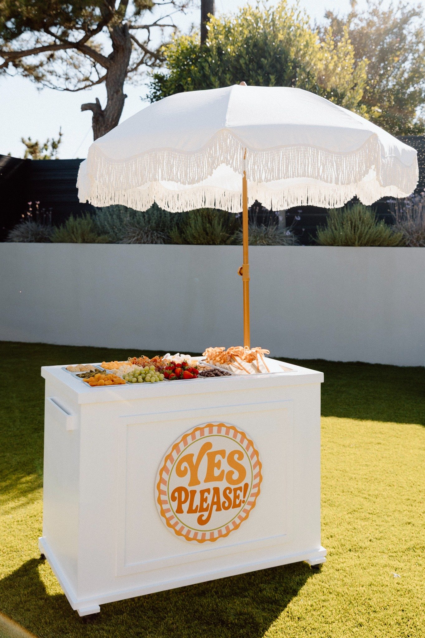 A white dessert table with a large umbrella on top, displaying a variety of fruits and snacks, with a logo that says "YES PLEASE!" on the front, set outdoors on grass during daytime.
