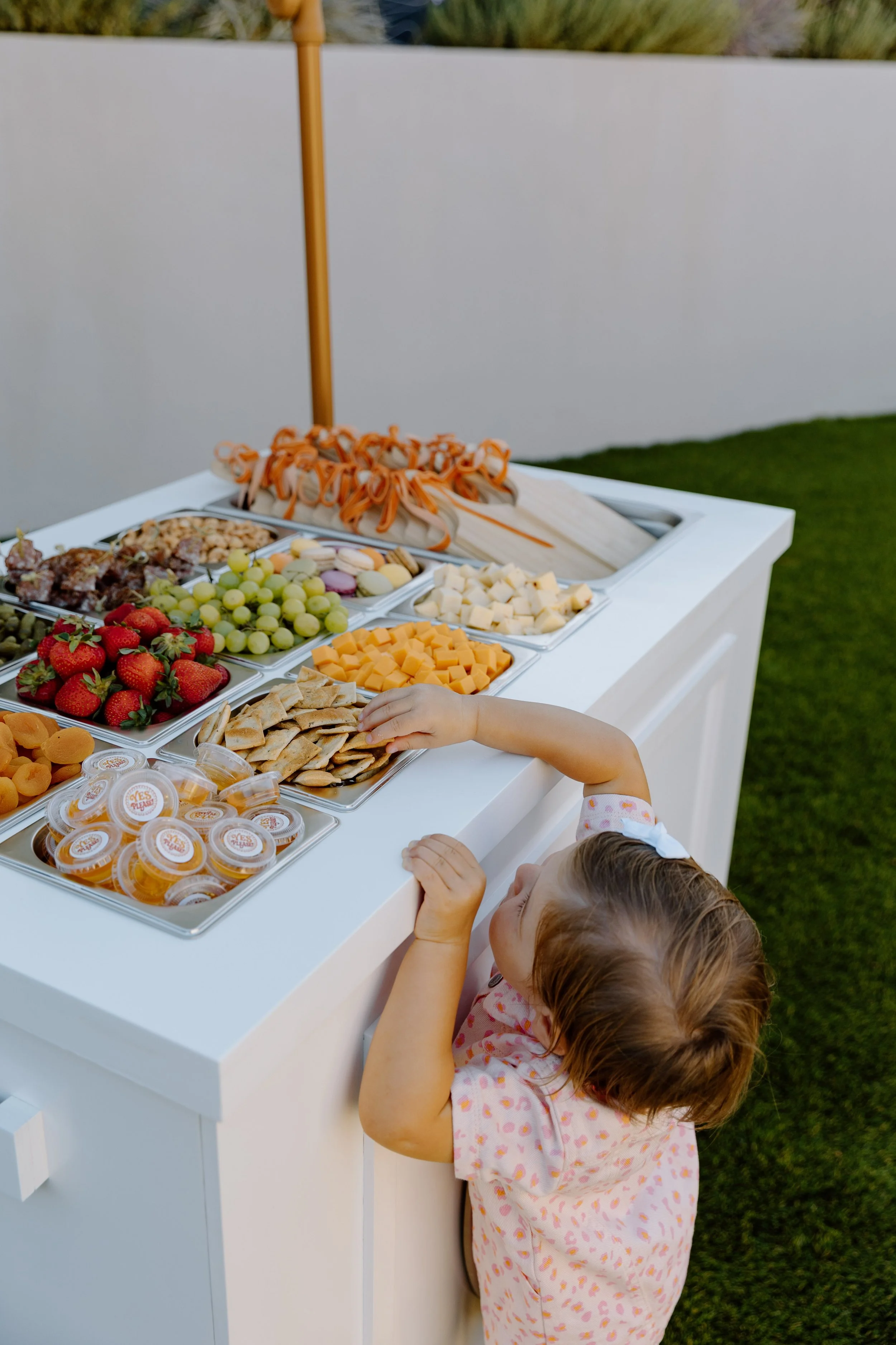 Close-up of a young girl reaching for food on a white buffet table outdoors, with various fruits, cheeses, and snacks arranged in trays, and a child's arm and head visible in the foreground.
