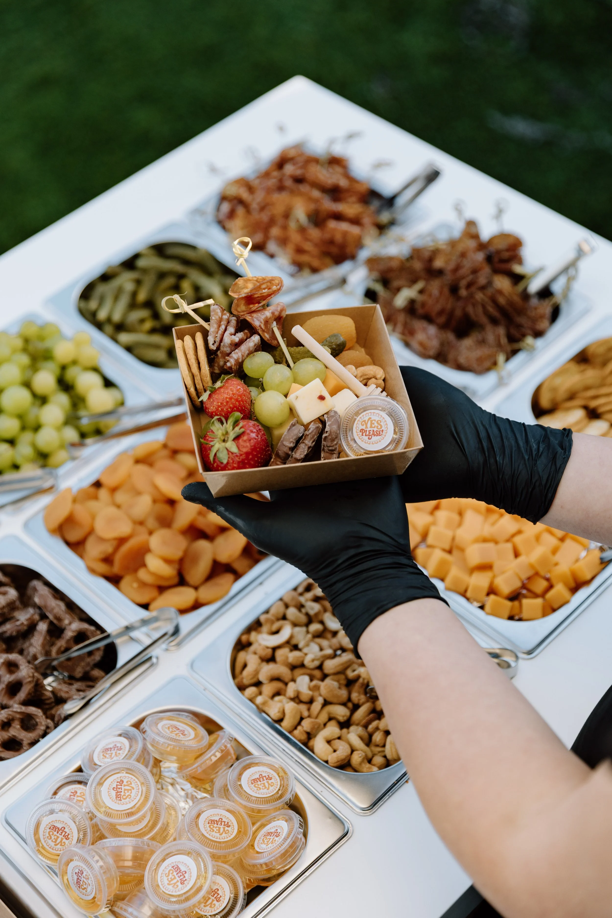 Person with black gloves holding a small tray of assorted snacks and cheese, with an assortment of cheeses, nuts, and fruits in the background.