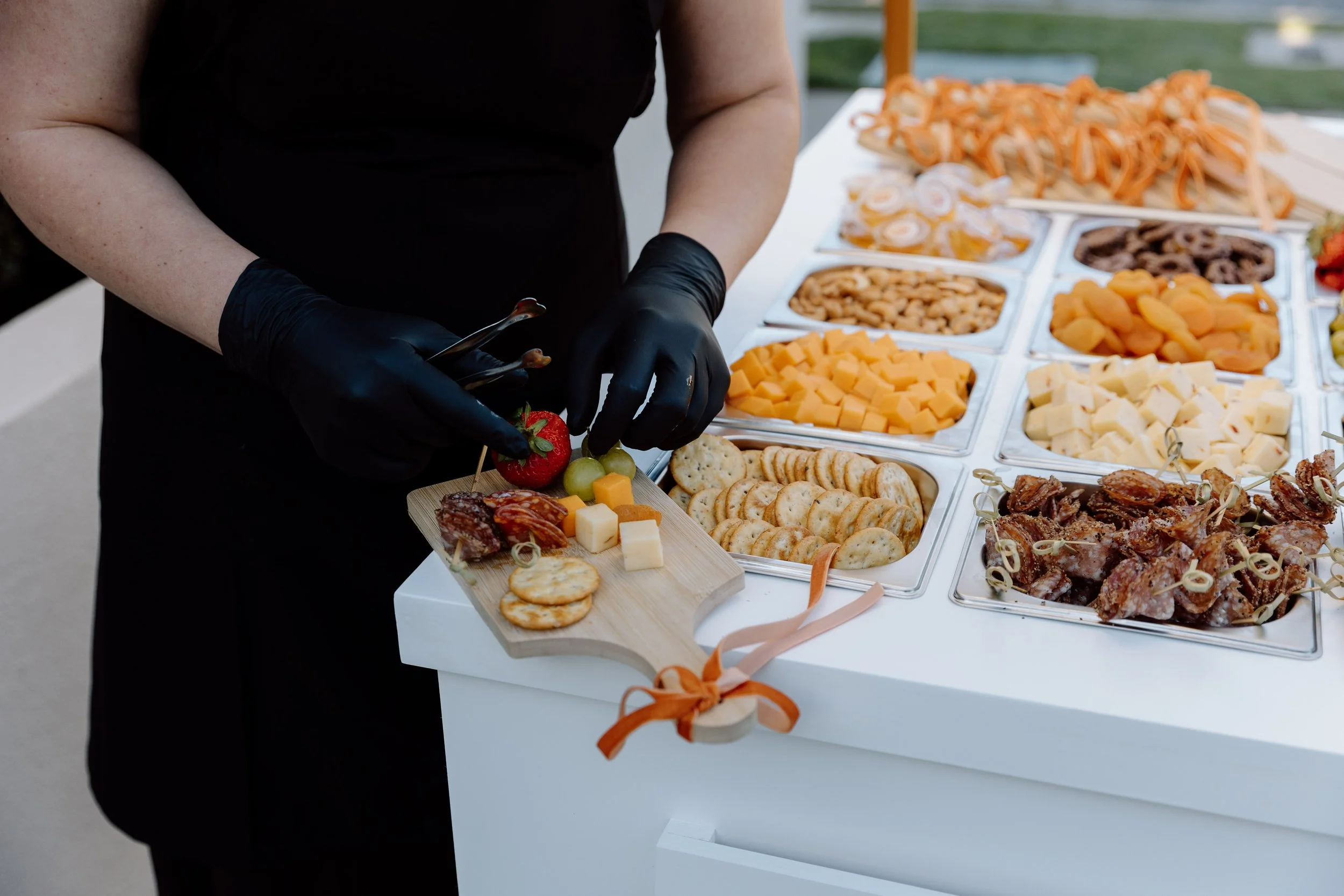 A person wearing black gloves prepares a platter of assorted cheeses, crackers, and fruits at an outdoor buffet table, with a table of various appetizers and snacks in the background.