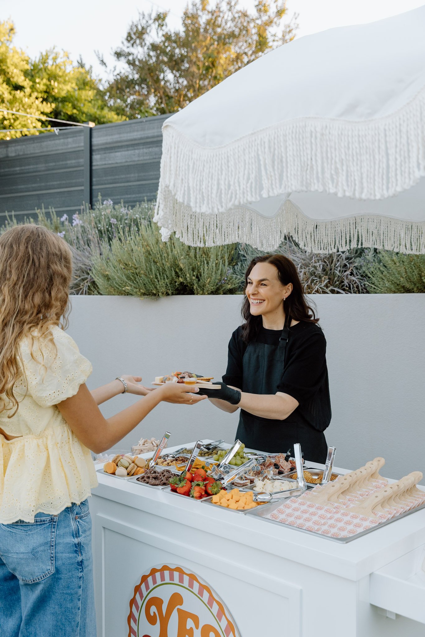 A woman in a black apron and gloves is serving food from a table with a variety of snacks, fruits, and desserts on it to a woman in a yellow dress with curly hair at an outdoor event.