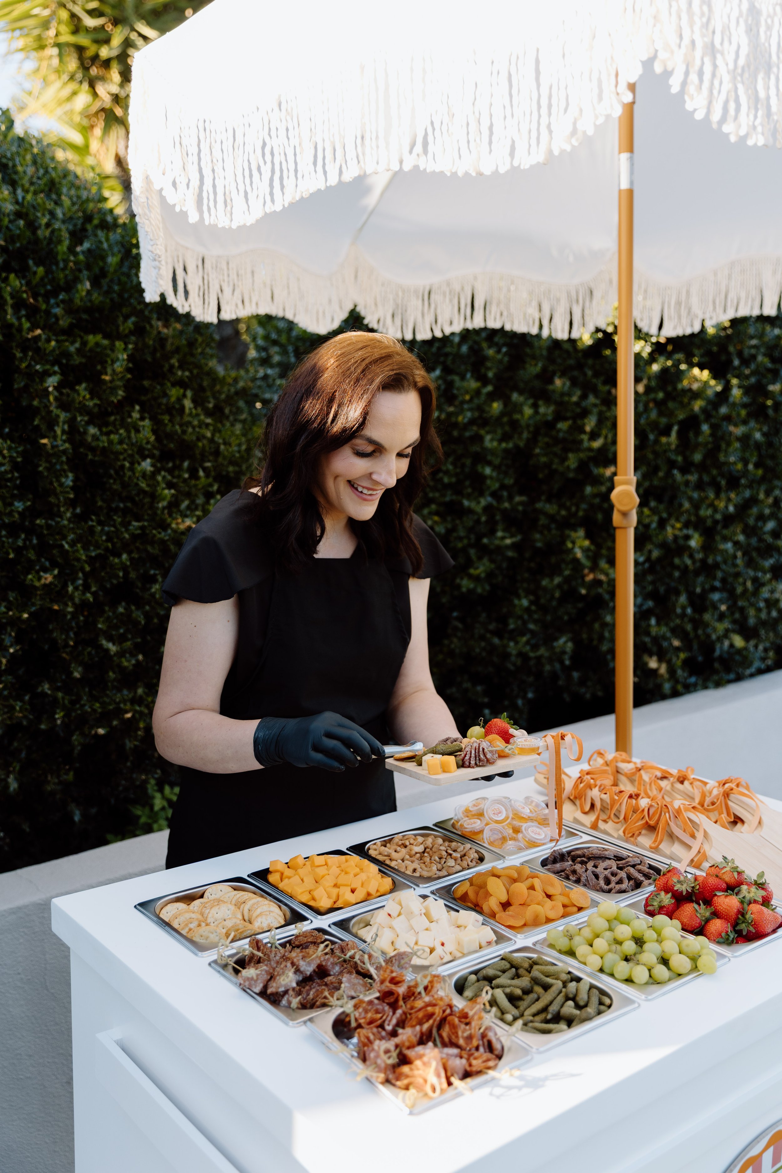 A woman with brown hair and a black dress serving a charcuterie platter at an outdoor buffet table under a white umbrella with fringes, surrounded by green bushes.
