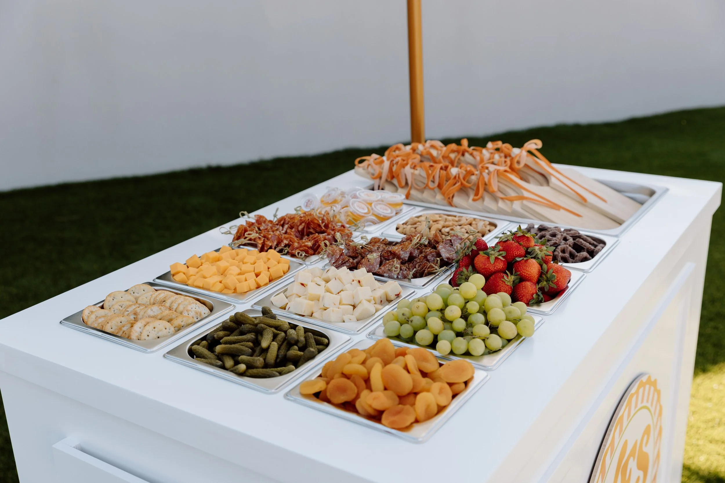 A white table with an assortment of cheeses, meats, fruits, vegetables, crackers, and snacks arranged in small metal trays for a buffet-style meal at an outdoor event.