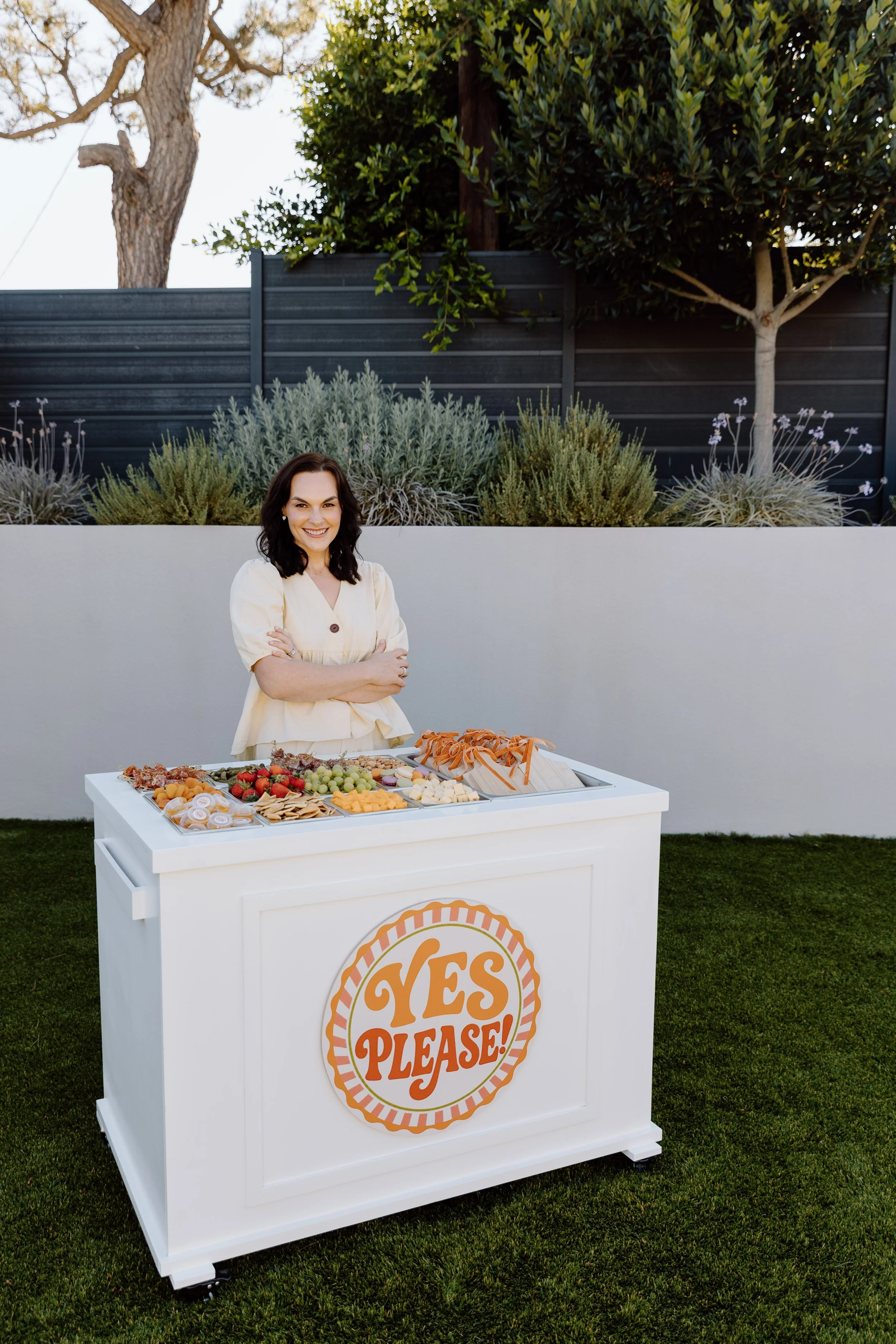 A woman with dark hair in a short bob style, wearing a light-colored dress, stands smiling with her arms crossed behind a white snack stand that has a sign reading "YES PLEASE!" in orange and white. The stand displays various snacks such as strawberries, grapes, cheese, crackers, and pretzels, set outdoors in front of a gray fence with greenery and trees in the background.