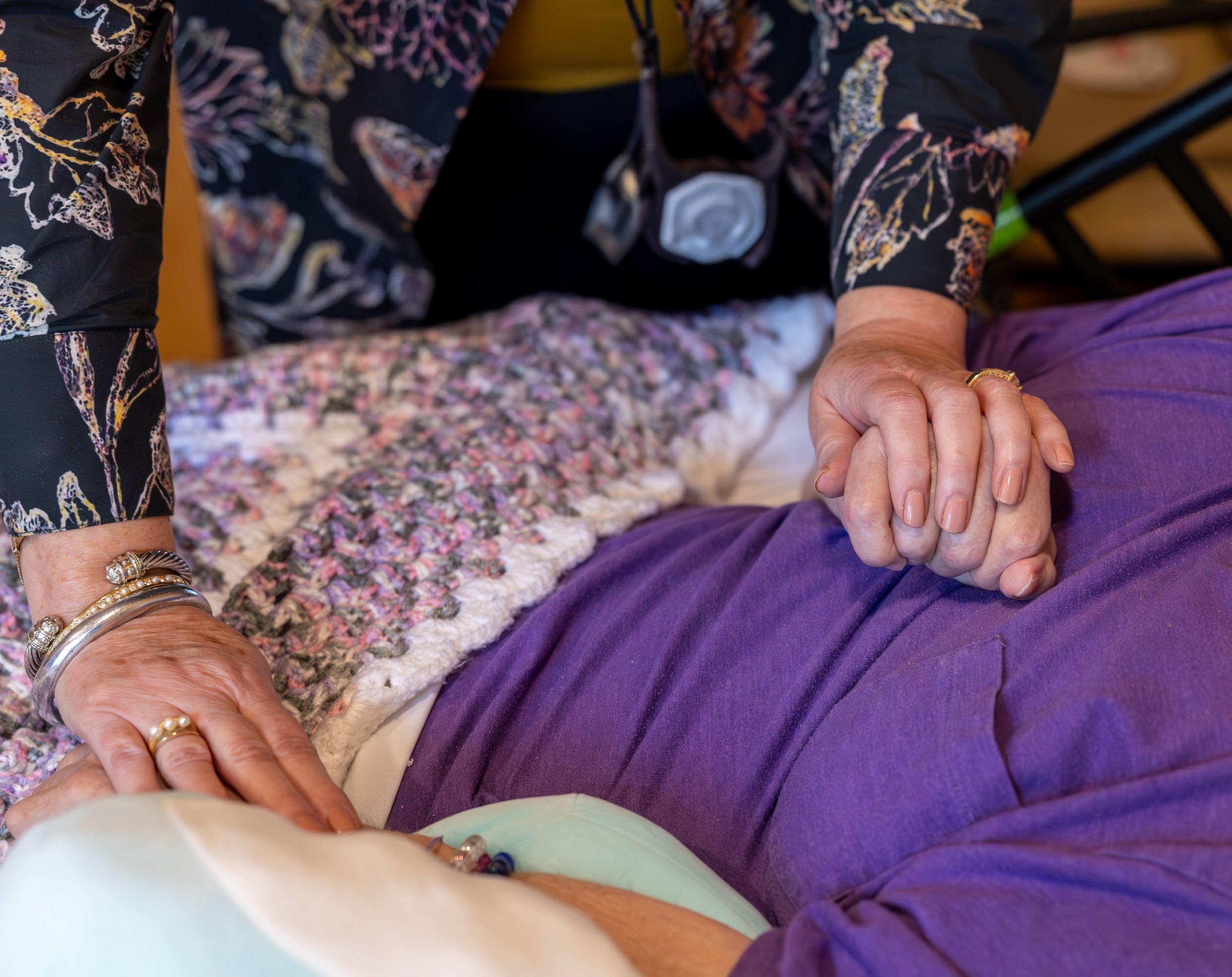Close-up of an elderly woman holding hands with a reiki practitioner, with both wearing rings. The elderly woman has jewelry on her wrists and is dressed in purple. In the background, there is a colorful, textured blanket.