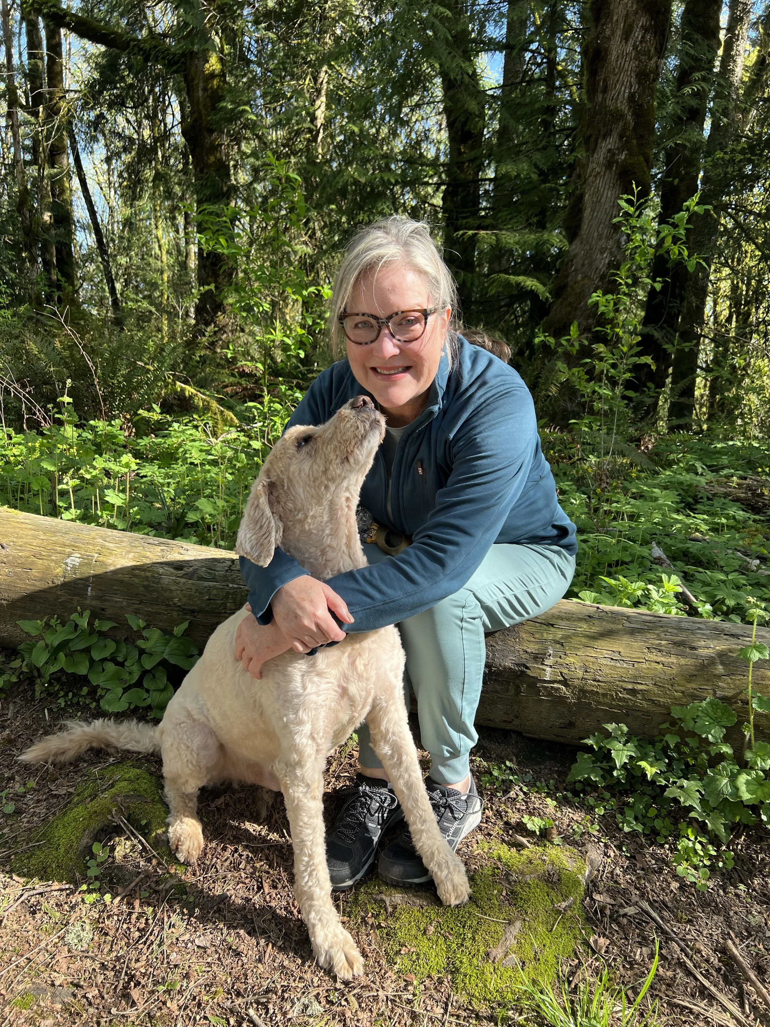 Theresa Trelstad wearing glasses and gray hair kneeling in a forest, hugging a large, light-colored dog with a floppy ear. The dog is standing on the ground with the woman. Sunlight filters through the tall trees in the background.