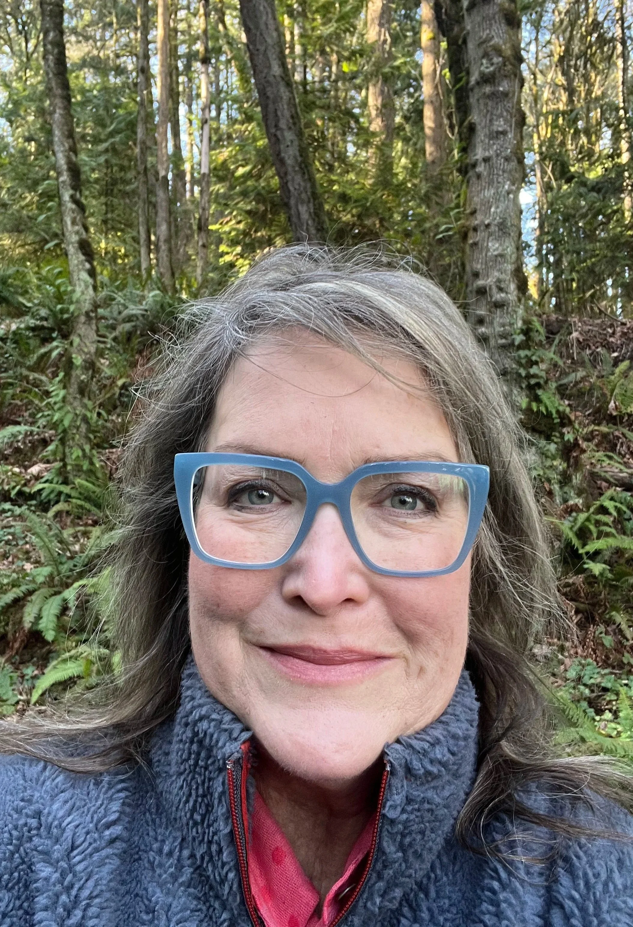 A woman with gray hair wearing blue glasses, a gray fleece jacket, and a pink scarf standing outdoors in a wooded area with trees and green foliage.
