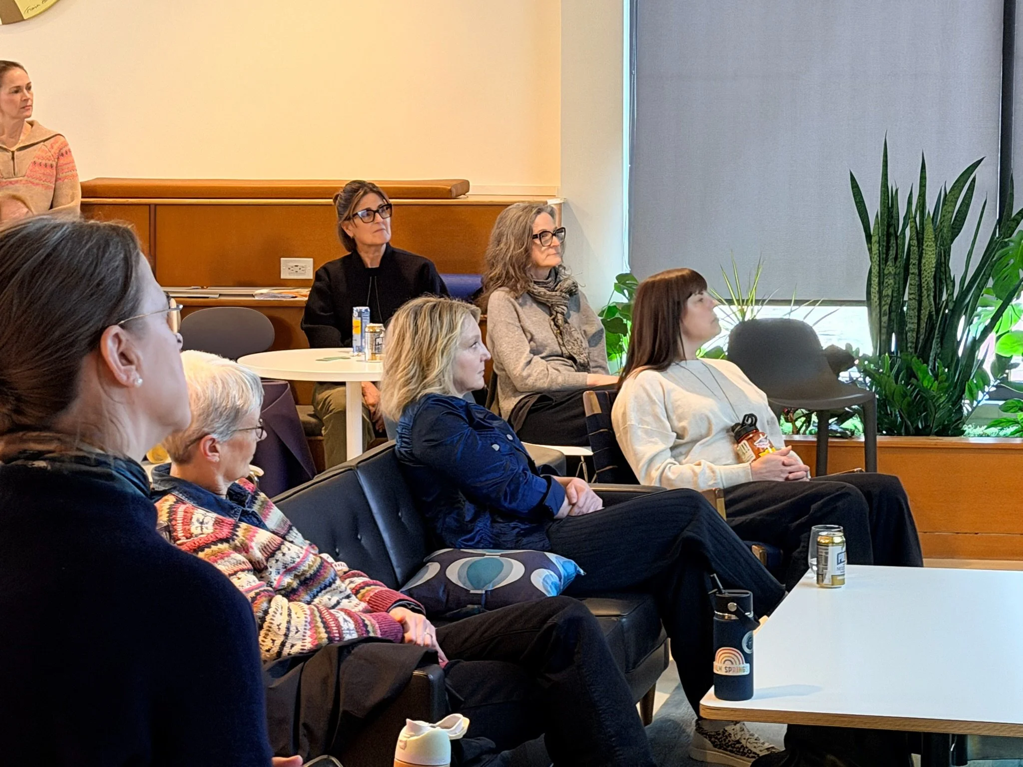 A group of women seated at tables, attentively listening to a presentation by Theresa Trelstad during a 1/2 Day, Multi-Modality Custom Retreat in a conference room with plants and a window.