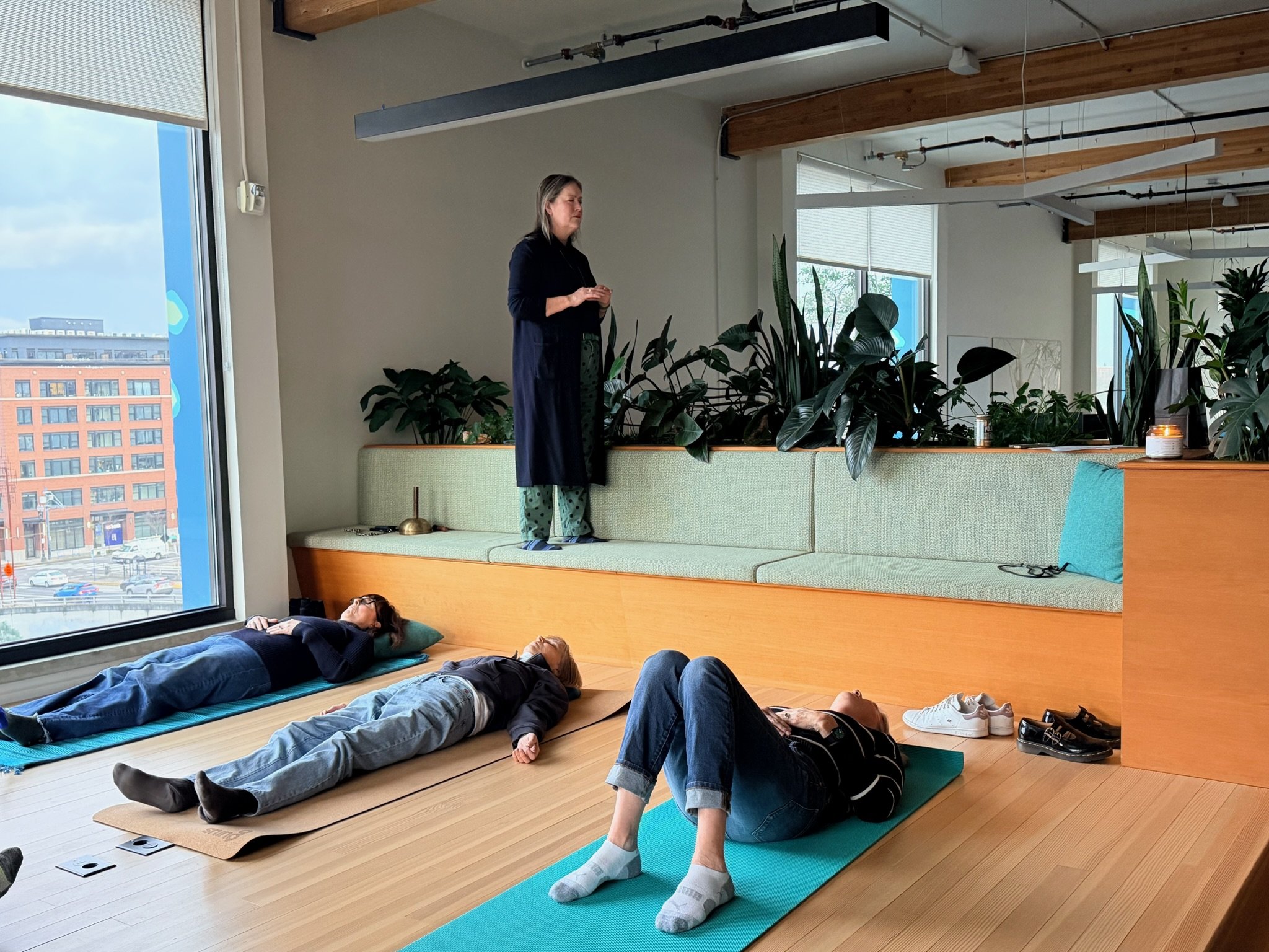 Theresa Trelstad with clients leading a Reiki Group Session in a bright, modern room with large windows, wooden ceiling beams, and an abundance of indoor plants.