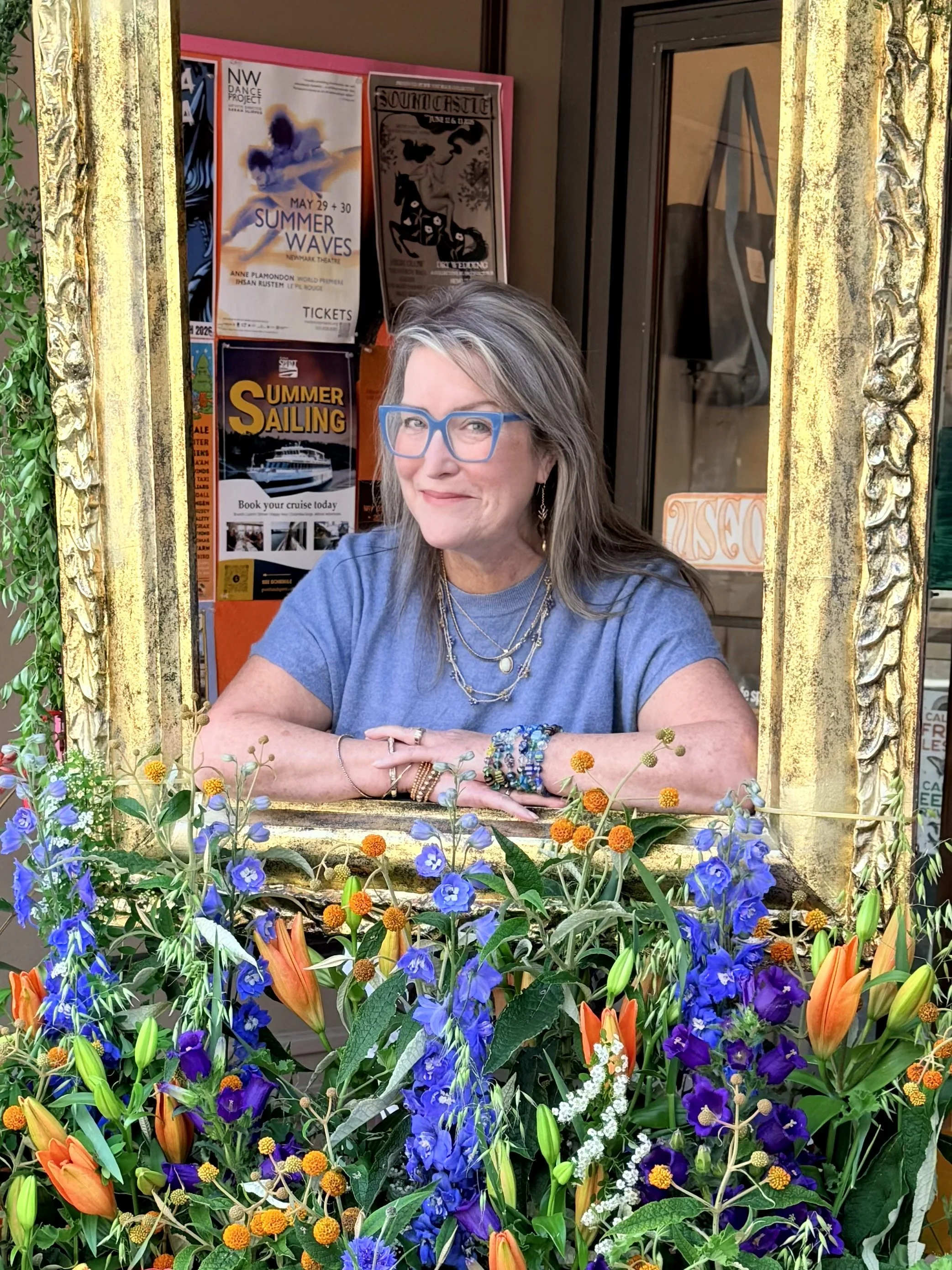 Theresa Trelstad wearing glasses sitting behind a decorative gold frame window, surrounded by a colorful arrangement of various flowers including orange lilies, blue and purple flowers, and green foliage.