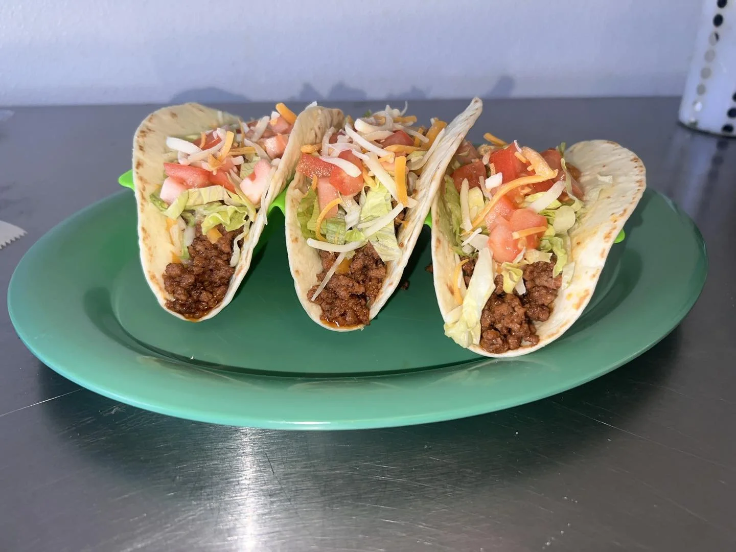 Three tacos with ground beef, lettuce, tomato, shredded cheese, and sour cream on a green plate.