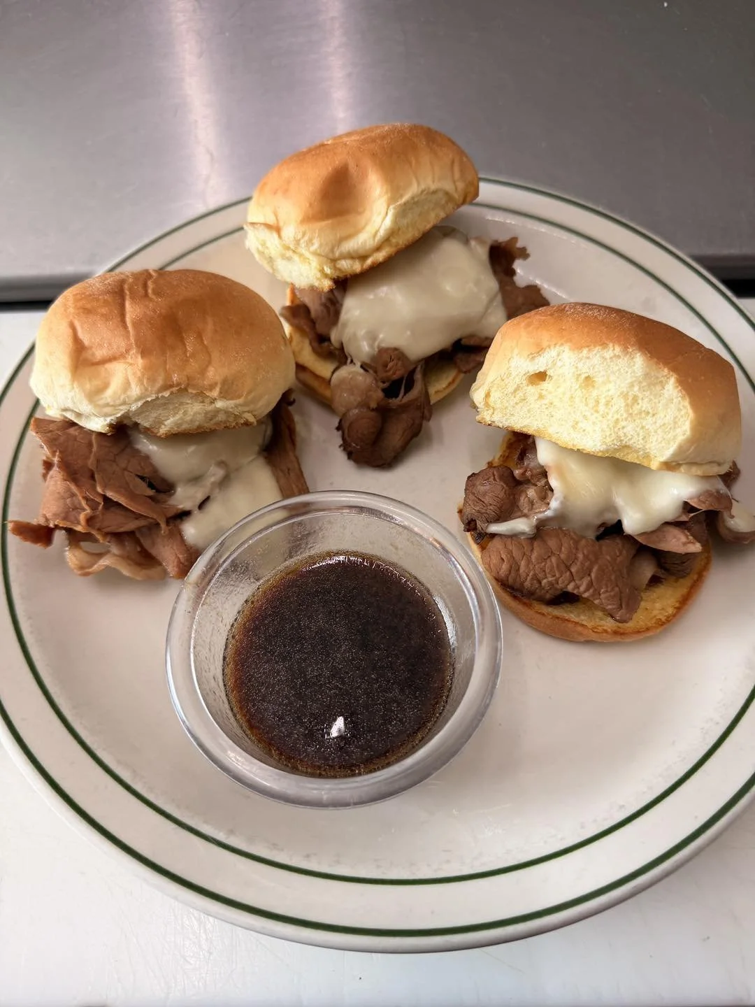 Three beef sliders with cheese on a plate, with a small cup of dark sauce.