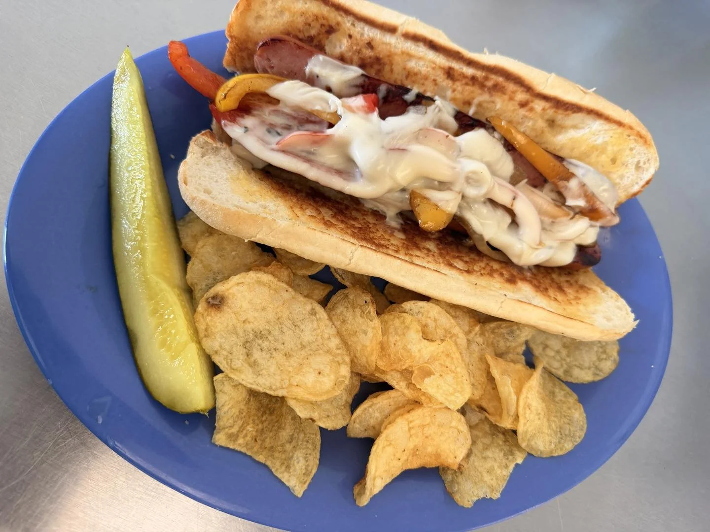 Hot dog with grilled bun, topped with relish, peppers, onions, and mayonnaise, served with potato chips and a pickle spear on a blue plate.