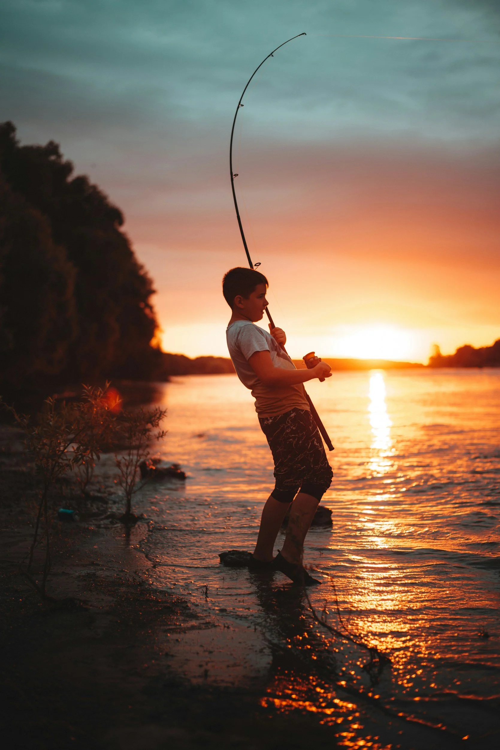 A boy fishing at sunset by a river, standing in shallow water with a fishing rod, silhouetted against the colorful sky.