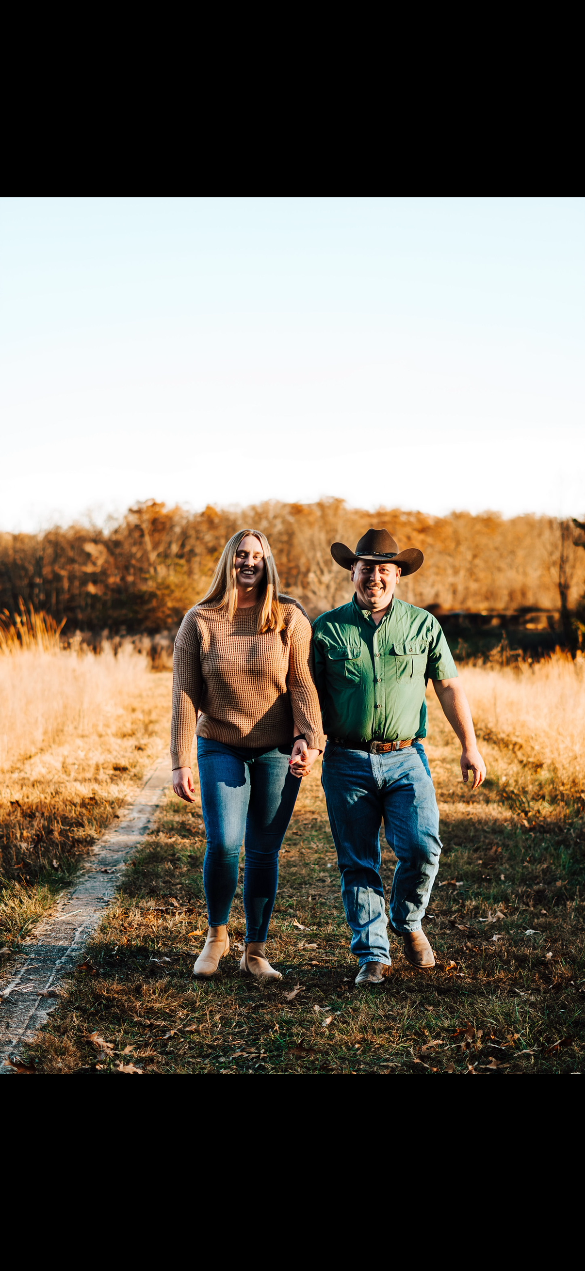 A smiling couple walking hand in hand outdoors on a dirt path through a field with tall grass and trees in the background during golden hour.