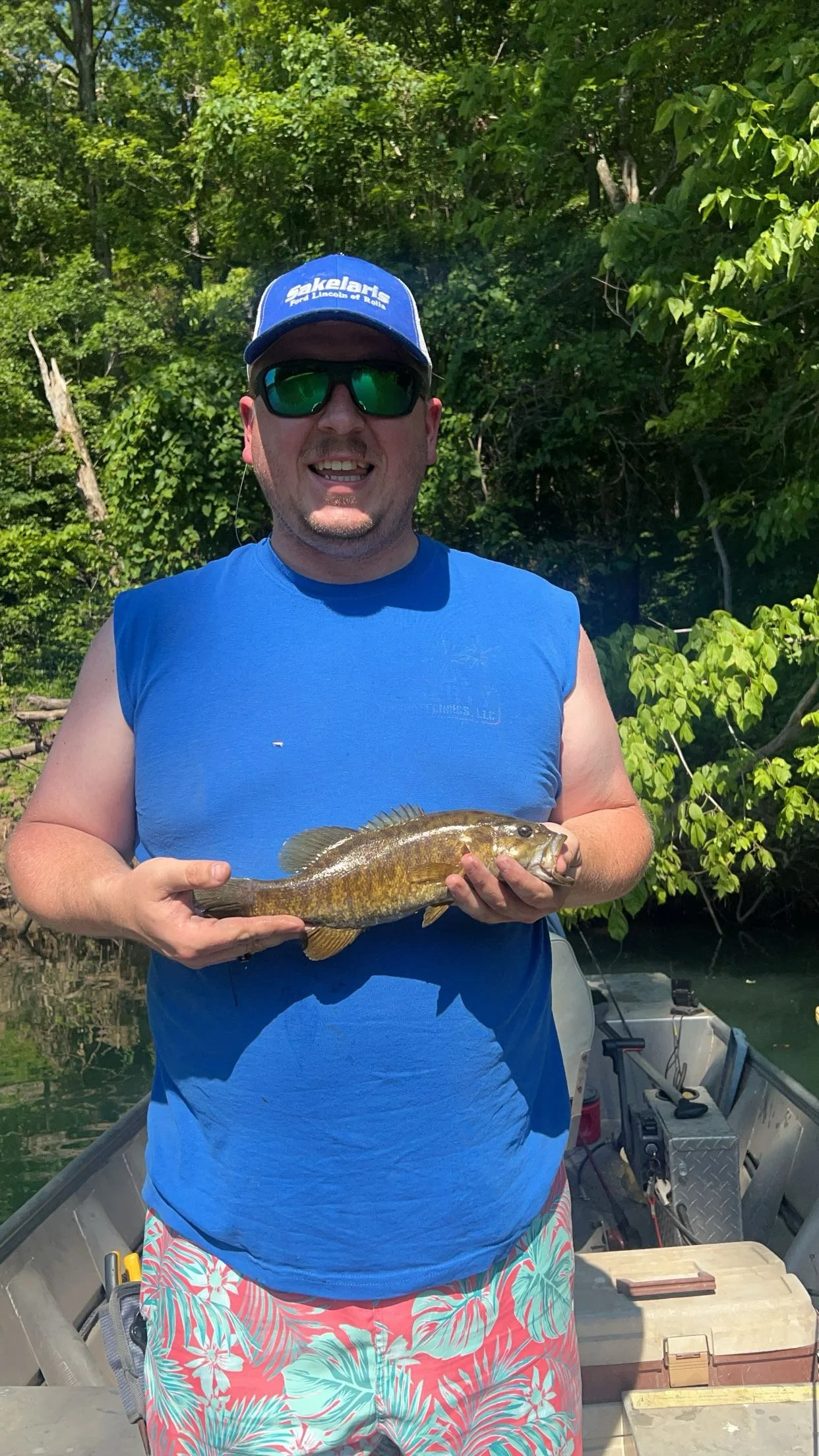 A man standing in a boat holding a fish, wearing sunglasses, a blue cap, a blue sleeveless shirt, and colorful shorts, with green foliage in the background.