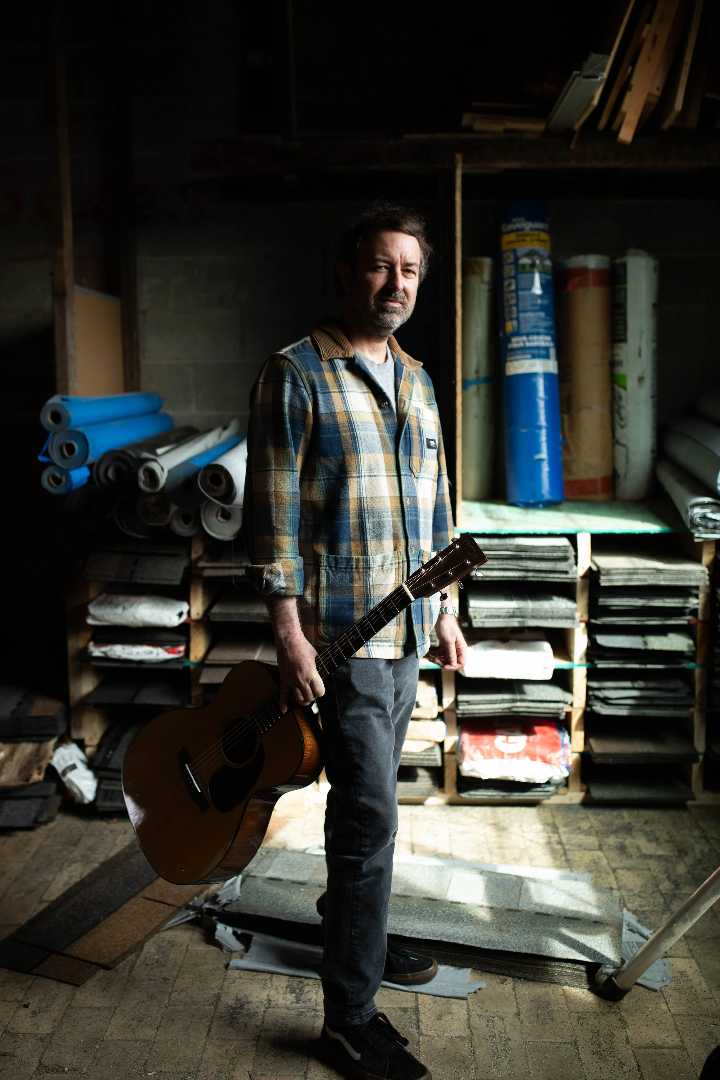 David Faul holding an acoustic guitar, standing in a workshop or storage room with shelves of rolled materials and various supplies behind him.