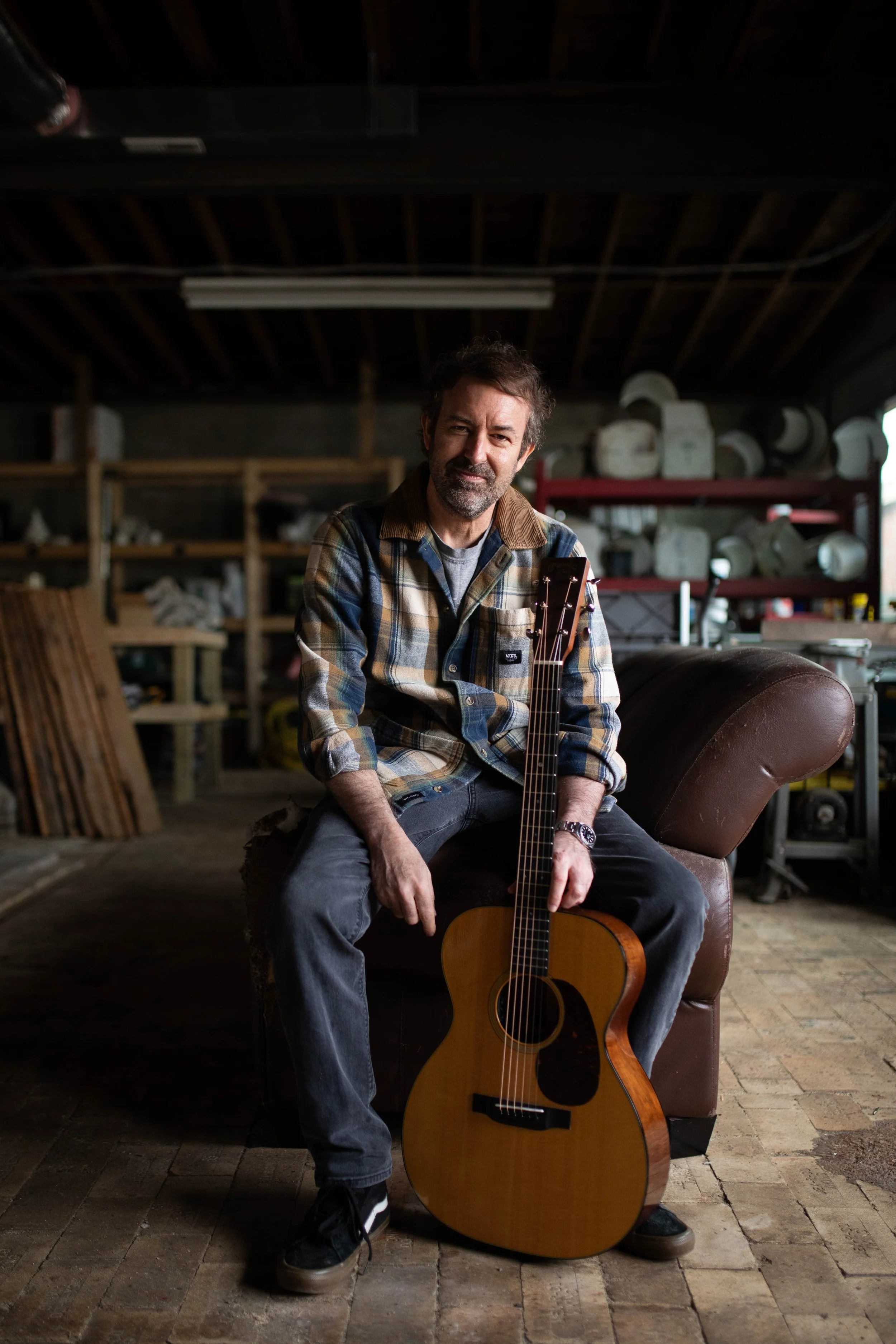 David Faul wearing a plaid shirt and jeans, sitting on a leather couch in a workshop or studio. He is holding an acoustic guitar, with shelves and various items in the background.