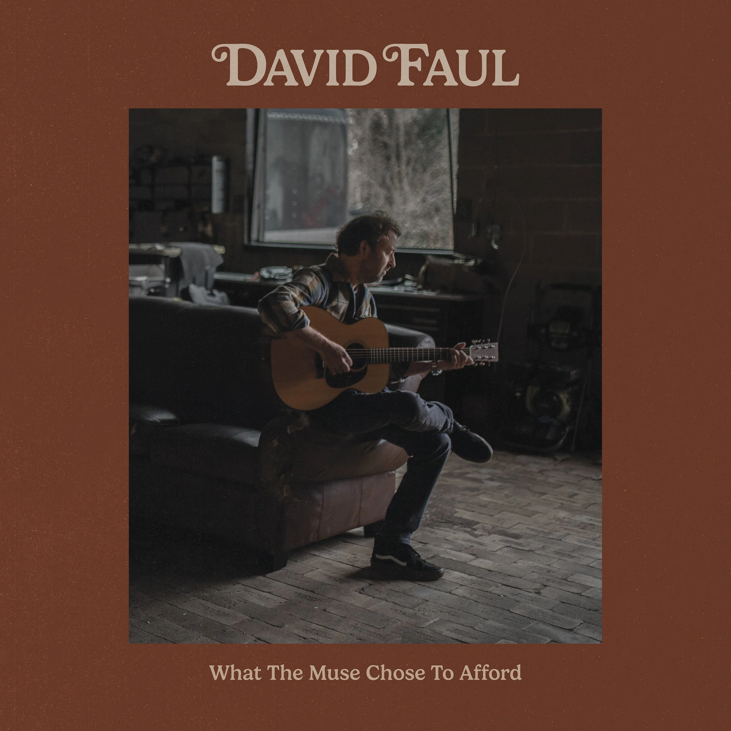 David Faul sitting on a couch playing an acoustic guitar inside a rustic room with a large window showing trees outside. The text overlay reads "What The Muse Chose To Afford".