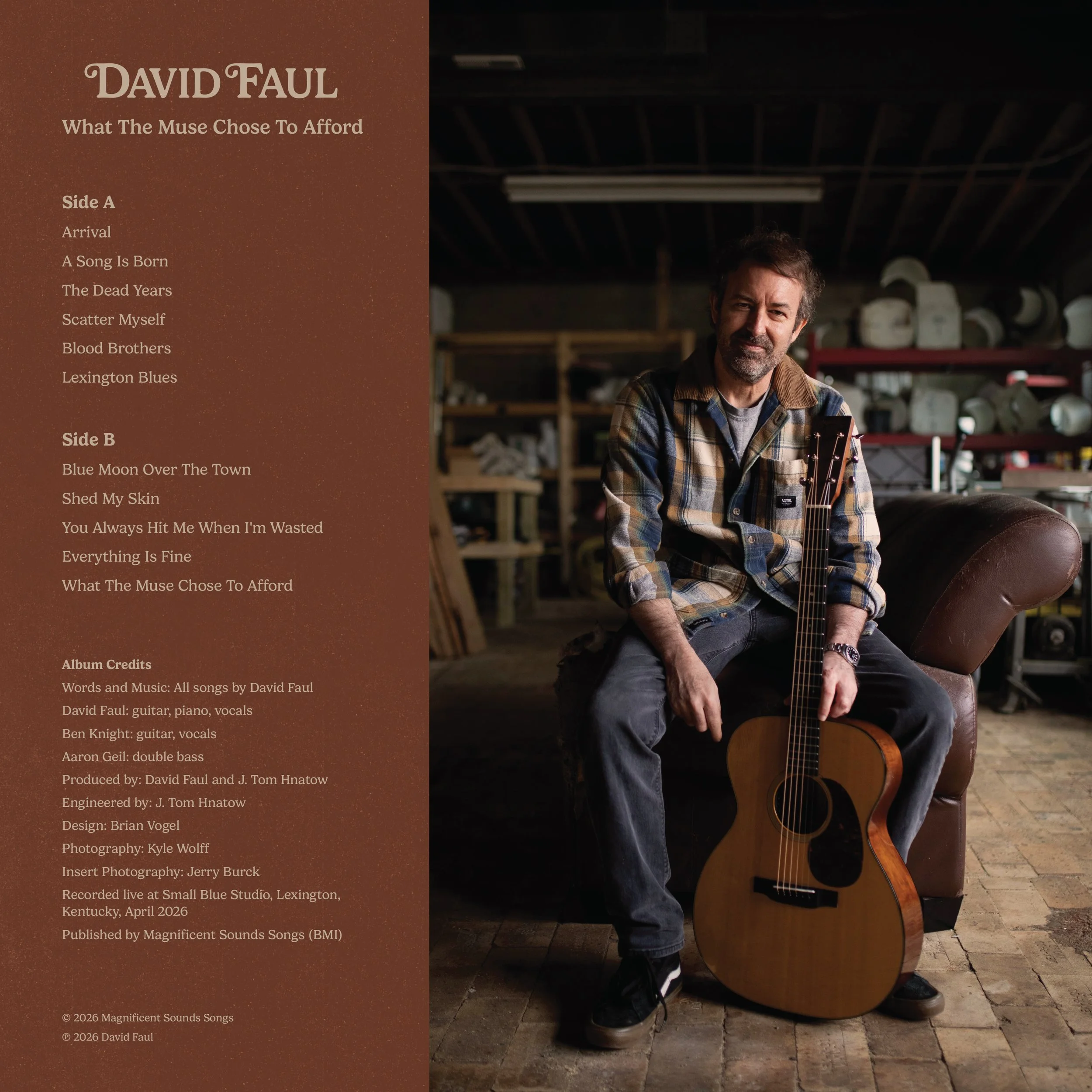 David Faul sitting in a warehouse, holding an acoustic guitar and smiling, with shelves of various objects in the background.